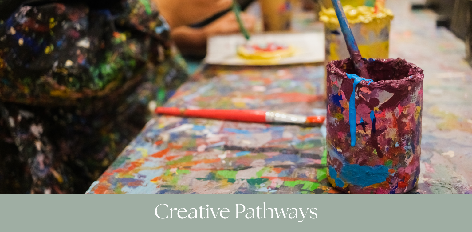 Painted cup with blue paint drips on a colorful, paint-stained table, with people painting in the background.
