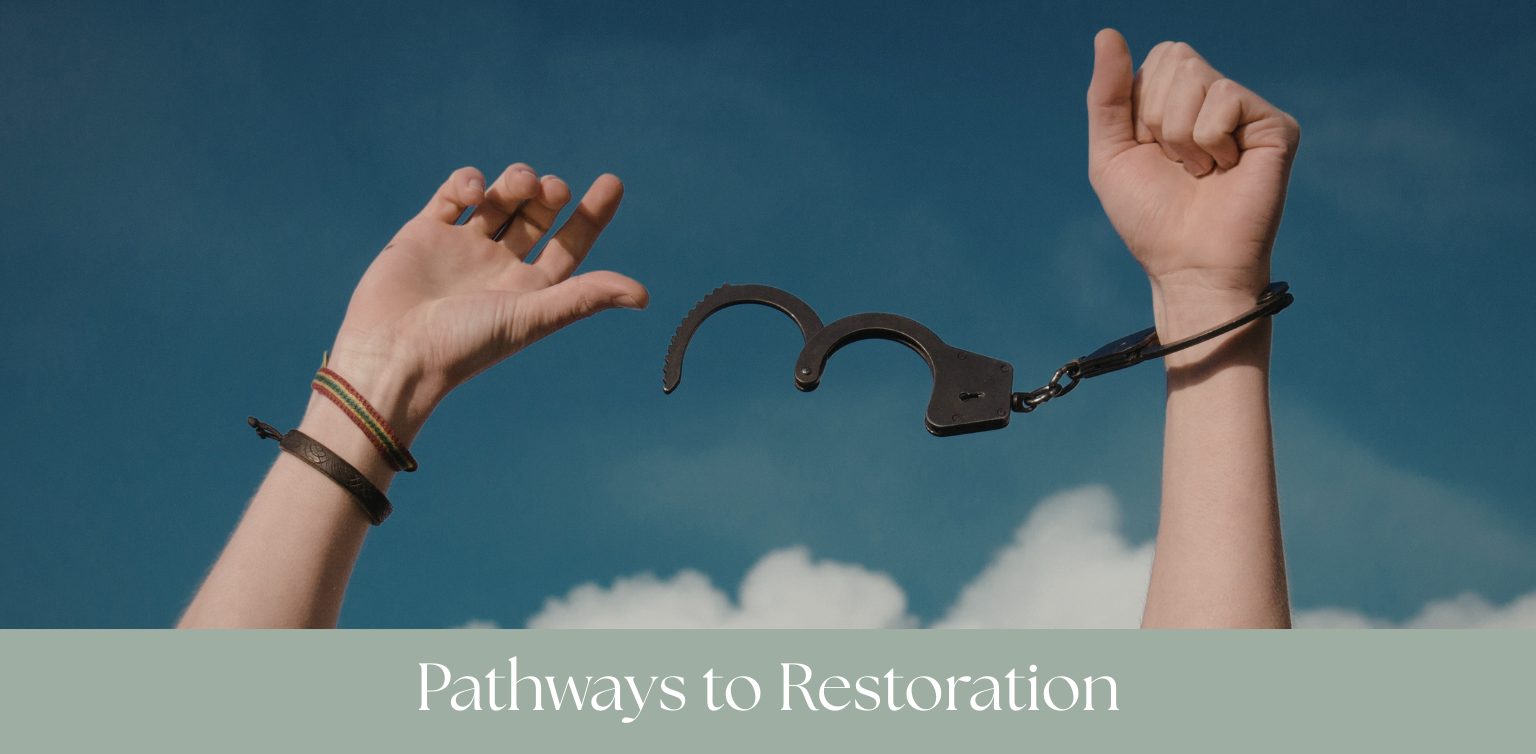 Two hands raised in the sky, one with a bracelet and the other with a wrist strap, holding a pair of handcuffs against a cloudy sky background, with the text 'Pathways to Restoration' at the bottom.