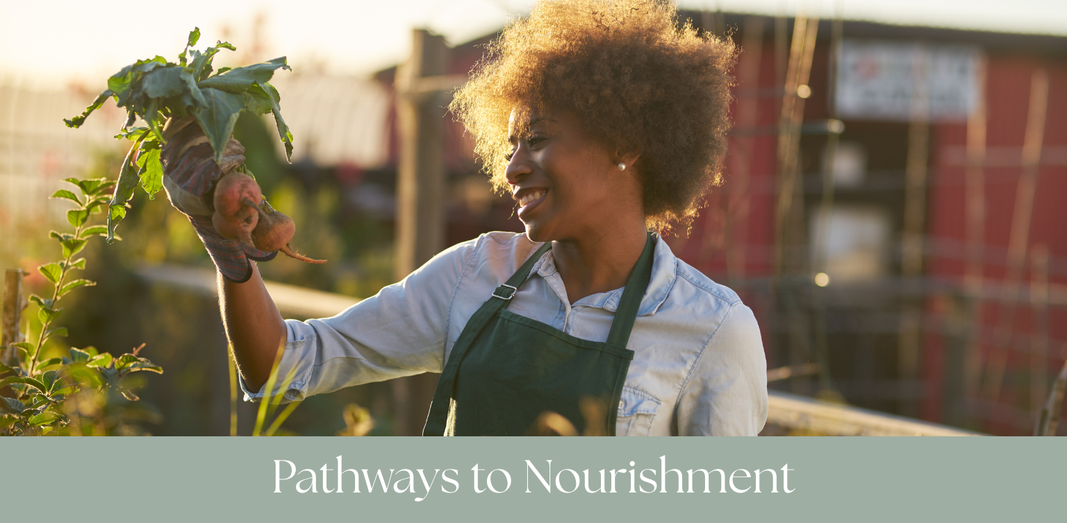 A woman with curly hair wearing a green apron and a denim shirt is harvesting sweet potatoes in a garden during sunset.