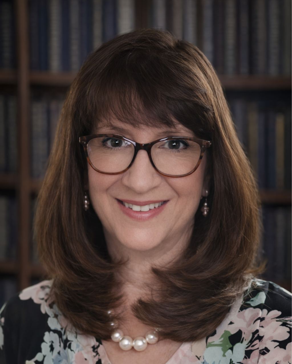 A woman with brown hair, glasses, and pearl jewelry smiling in front of a background of bookshelves filled with books.