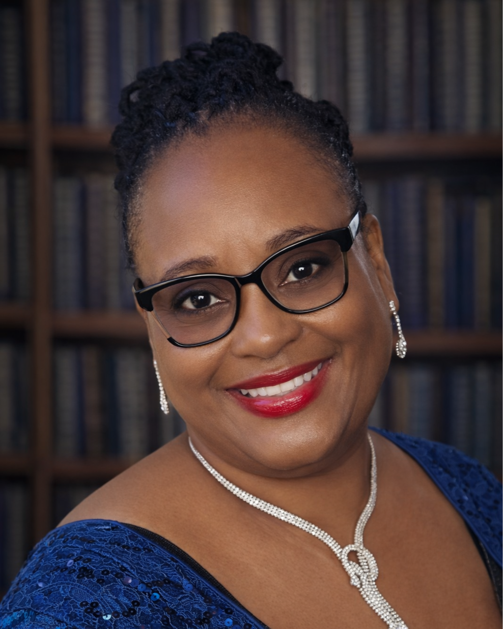 A smiling woman with glasses, diamond earrings, a diamond necklace, wearing a blue dress with lace detailing, standing in front of a bookshelf filled with books.