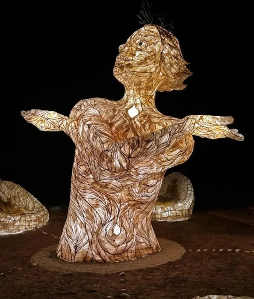 A large, illuminated sculpture resembling a mythical creature or dancer with arms outstretched, made of intertwined materials glowing from within against a dark background.