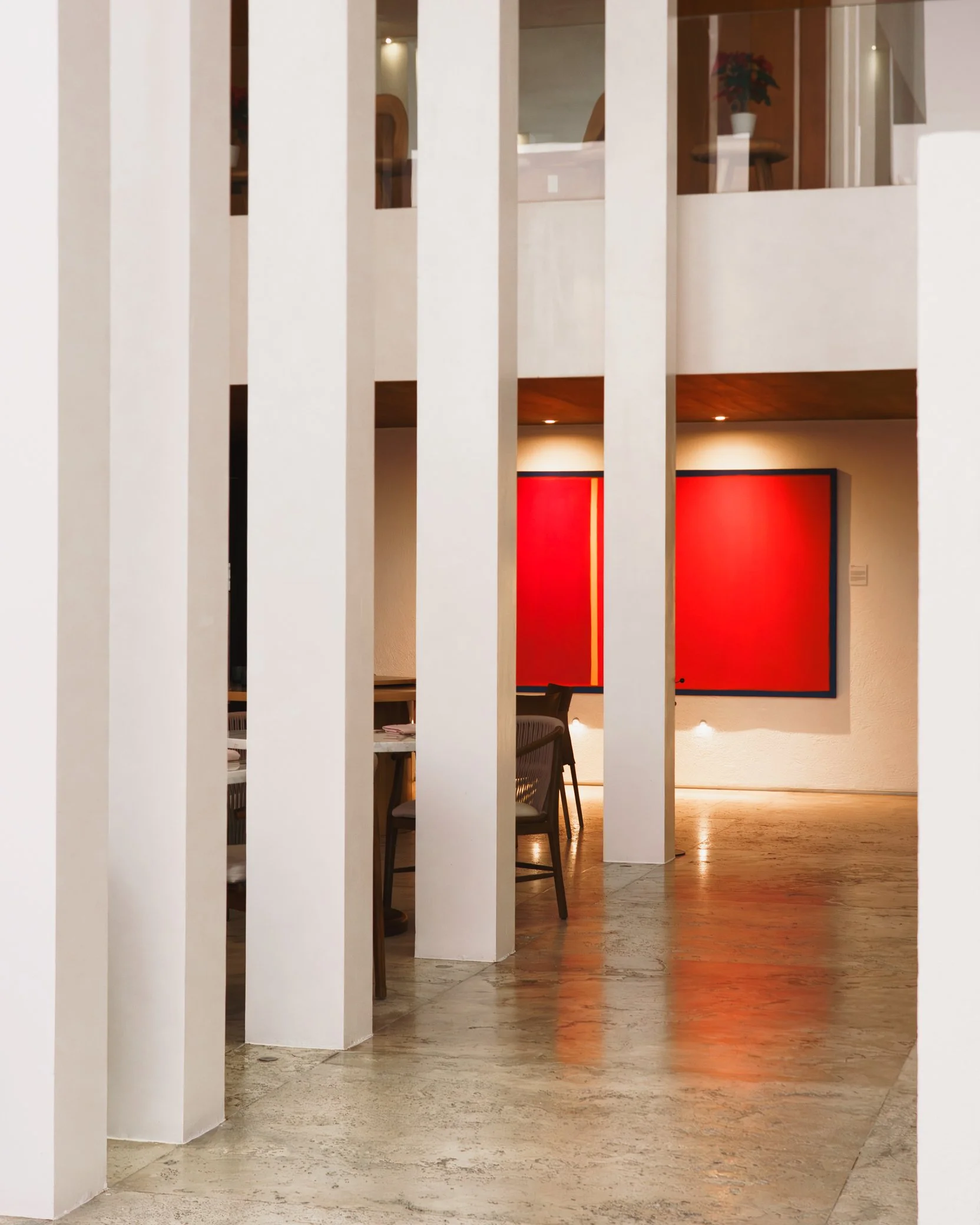 Interior of a modern building with vertical white wall dividers, wooden flooring, and a wall with a large red artwork. Visible furniture includes chairs and tables, with a potted plant on a table in the background.