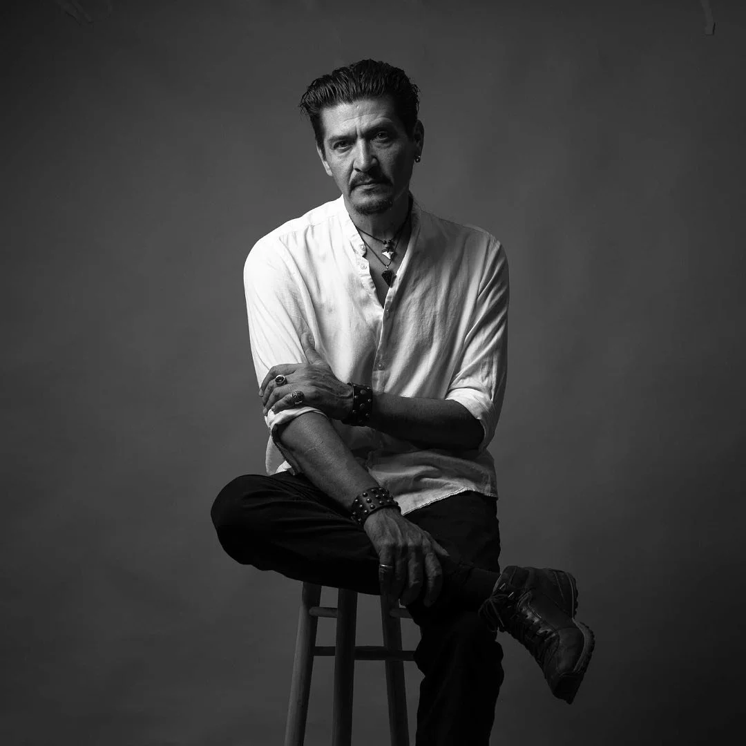 Black and white portrait of a man sitting on a stool, wearing a white shirt, dark pants, and black shoes, with various jewelry and accessories, against a plain background.