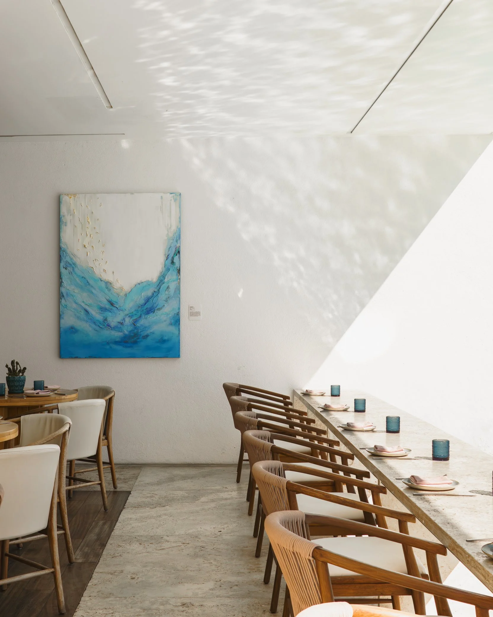 A modern dining area with a long, light-colored stone table set with plates, napkins, and blue glasses. Wooden chairs with rattan backs are positioned around the table, and additional chairs are visible on the other side. The wall features a large ab