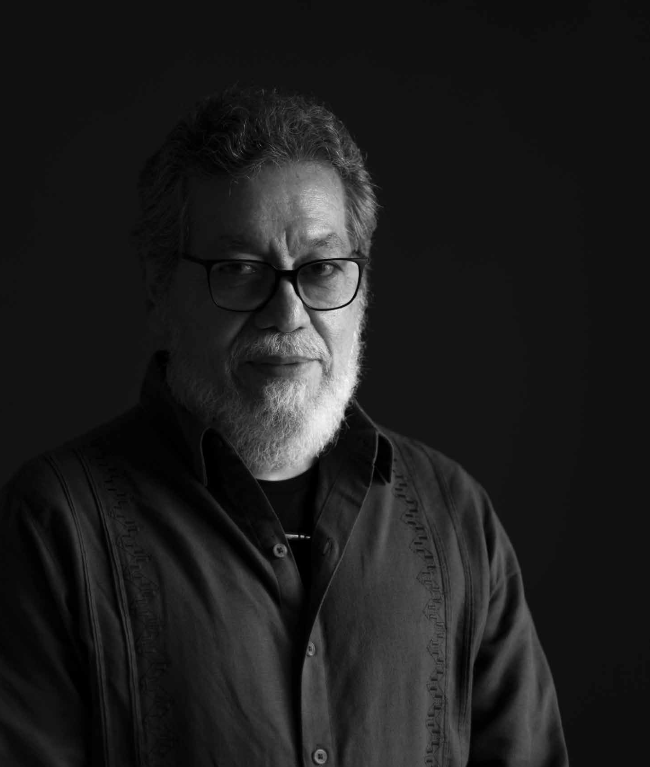 Black and white portrait of a middle-aged man with glasses, gray hair, and a beard, wearing a dark shirt against a dark background.