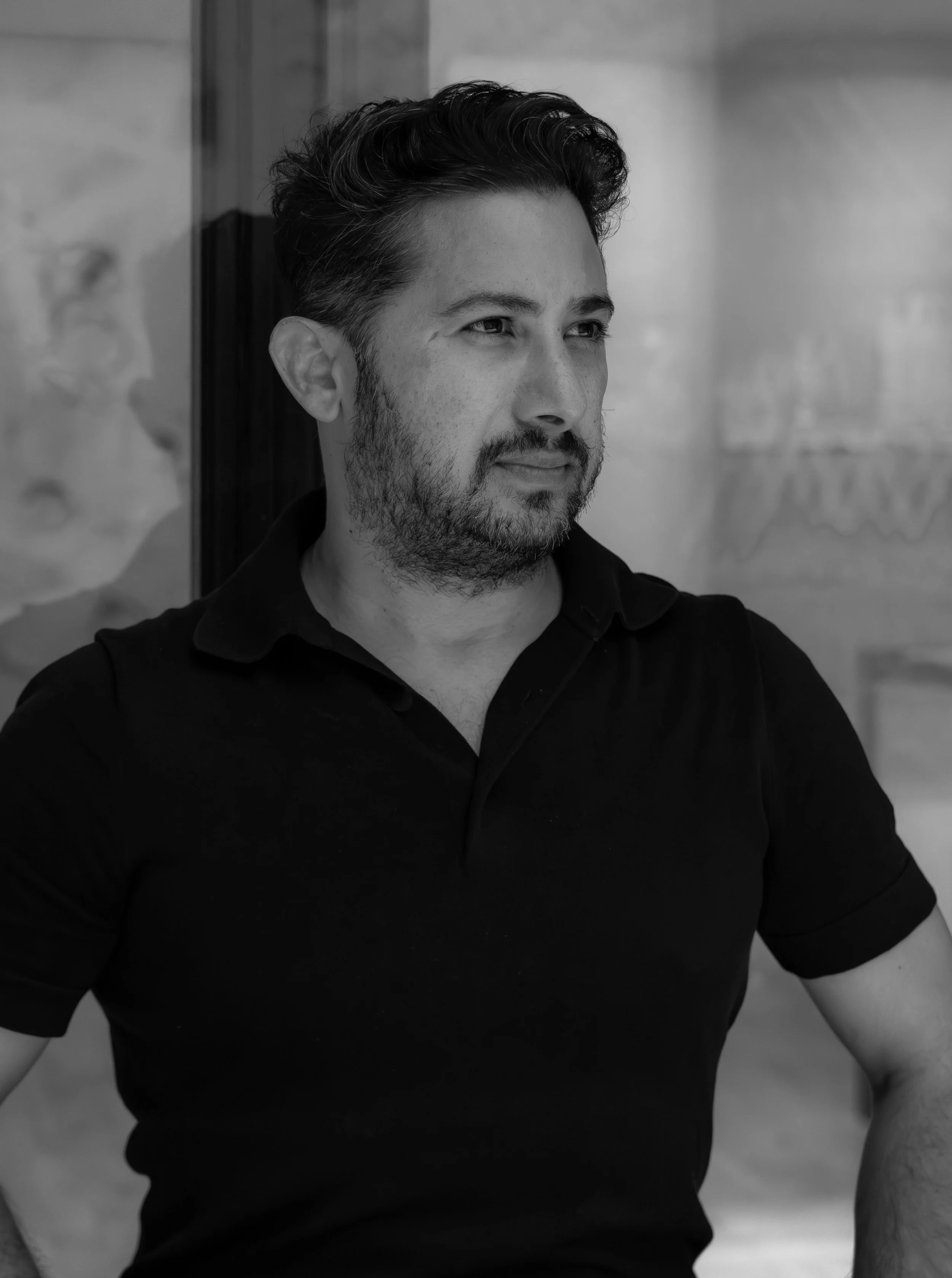 A man with dark, wavy hair and a beard, wearing a black collared shirt, looking to his right in front of a blurred background.