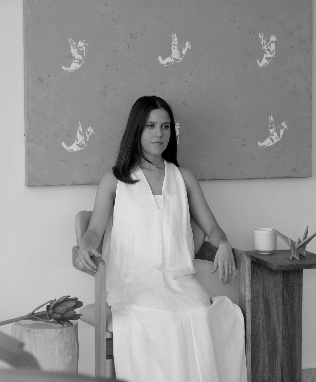 A woman with long dark hair sitting on a wooden chair in a white sleeveless dress in front of a textured wall with abstract artwork. A small wooden table with a white cup, a folded paper crane, and a decorative object is next to her, and a tulip flower is resting on a piece of wood in the foreground.