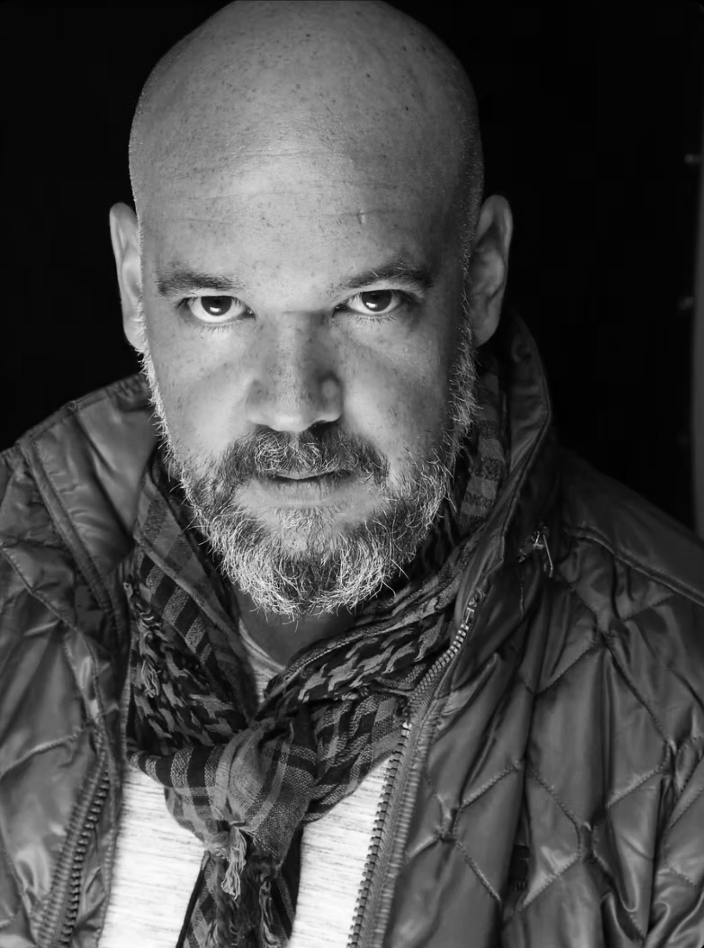Black and white close-up portrait of a bald man with a beard, wearing a quilted jacket and a scarf.