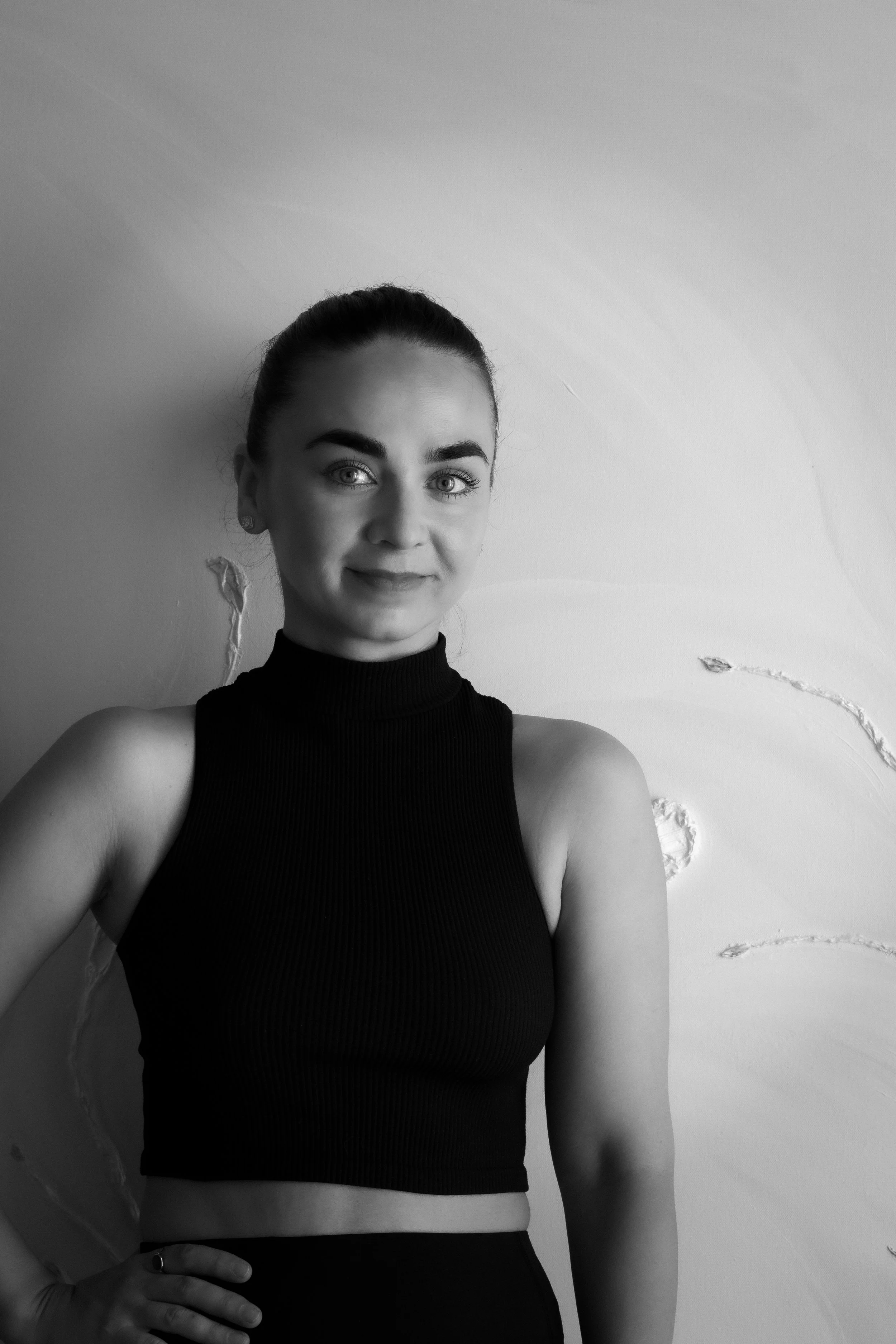 Black and white photo of a woman with dark hair tied back, wearing a sleeveless black top, posing with one hand on her hip against a textured wall.