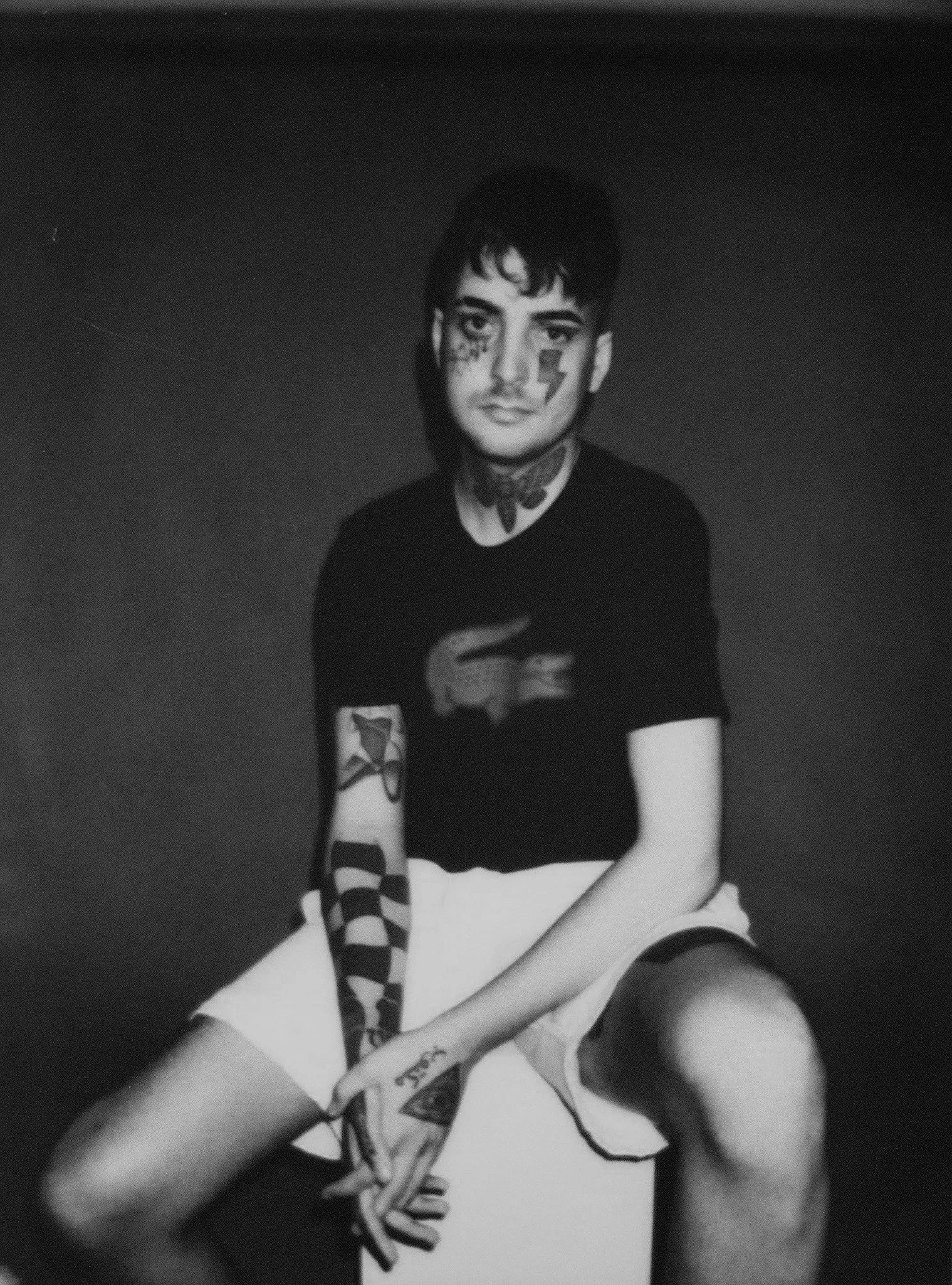 Black and white photo of a young person with tattoos and face paint, sitting with legs crossed, wearing striped socks and a black shirt, against a plain background.