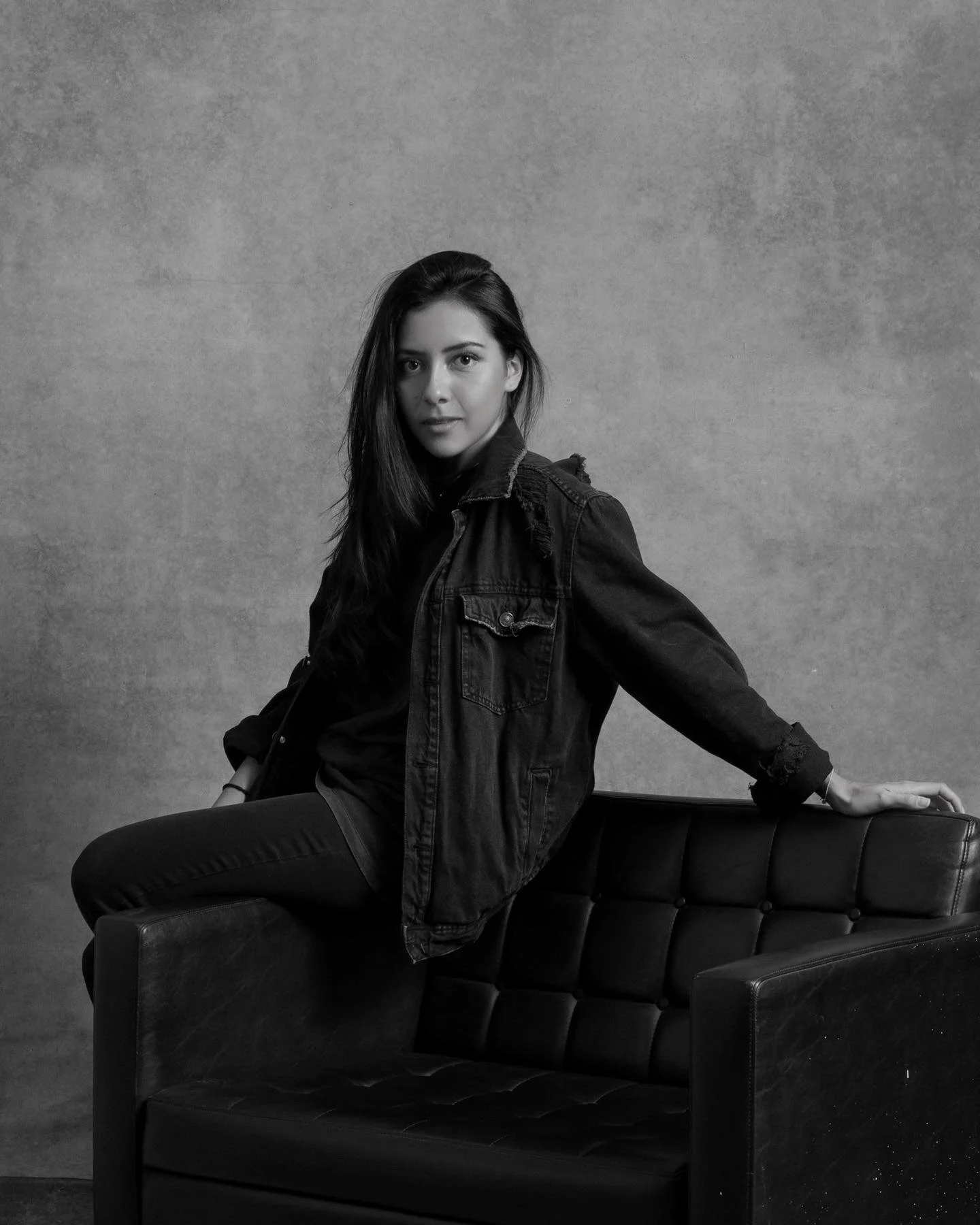 A young woman with dark hair sitting on the armrest of a black leather chair, wearing a denim jacket and looking at the camera against a plain textured background.