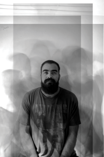 A man with a beard and mustache standing in front of a plain wall, with multiple ghostly copies of his image creating a blurred or motion effect.