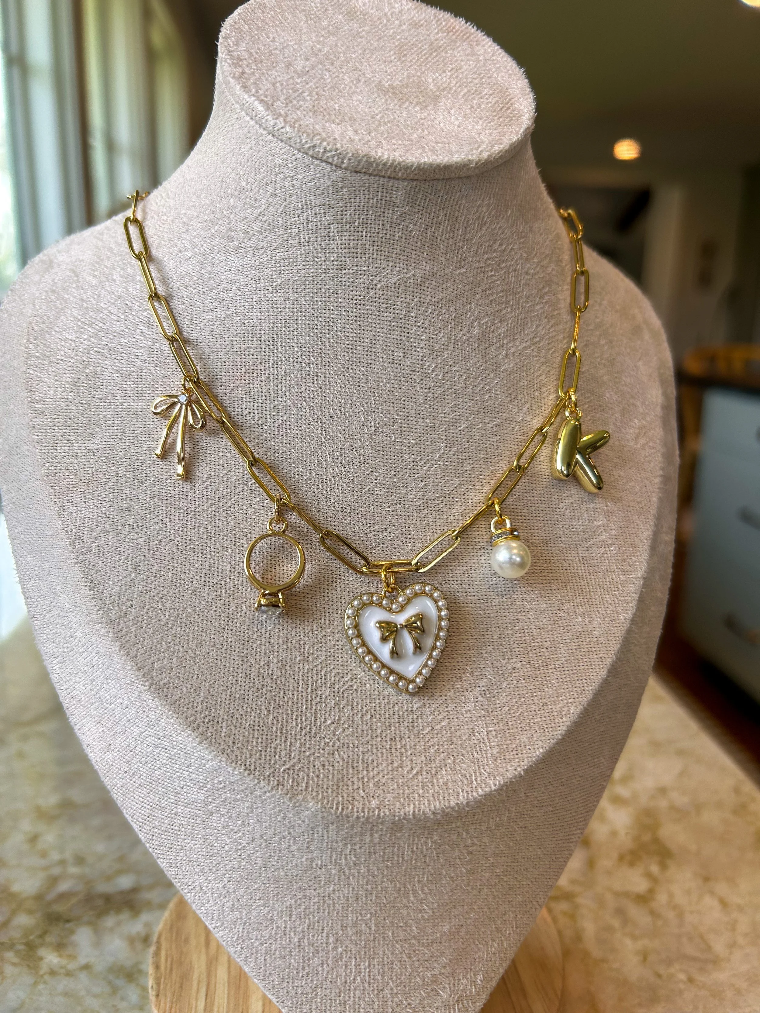 Gold chain necklace with charms including a bow, a ring, a pearl, a butterfly, and a heart with a bow design, displayed on a beige jewelry bust.
