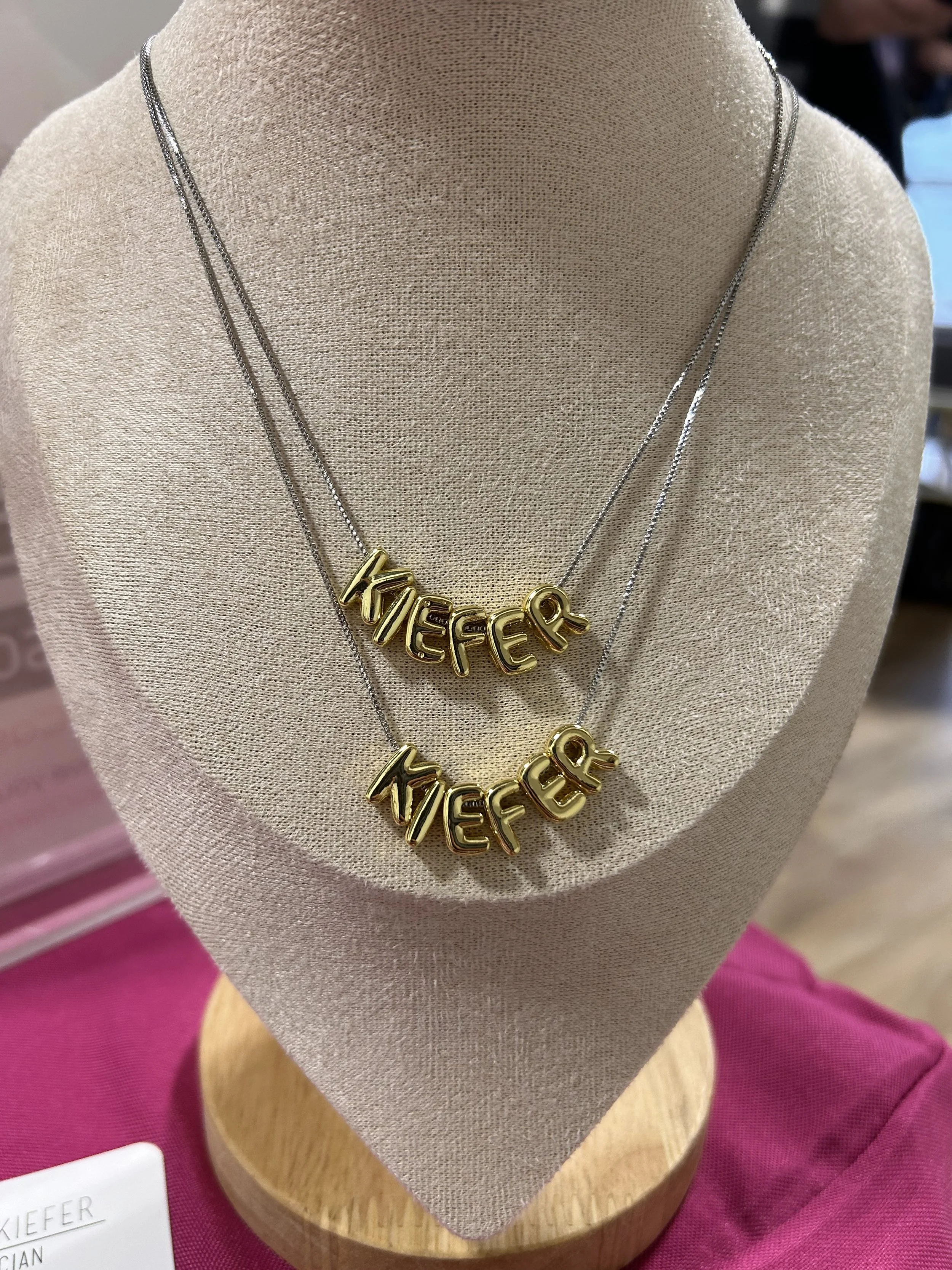 Two gold necklaces with the word 'KIEFER' in stylized letters on a display bust.