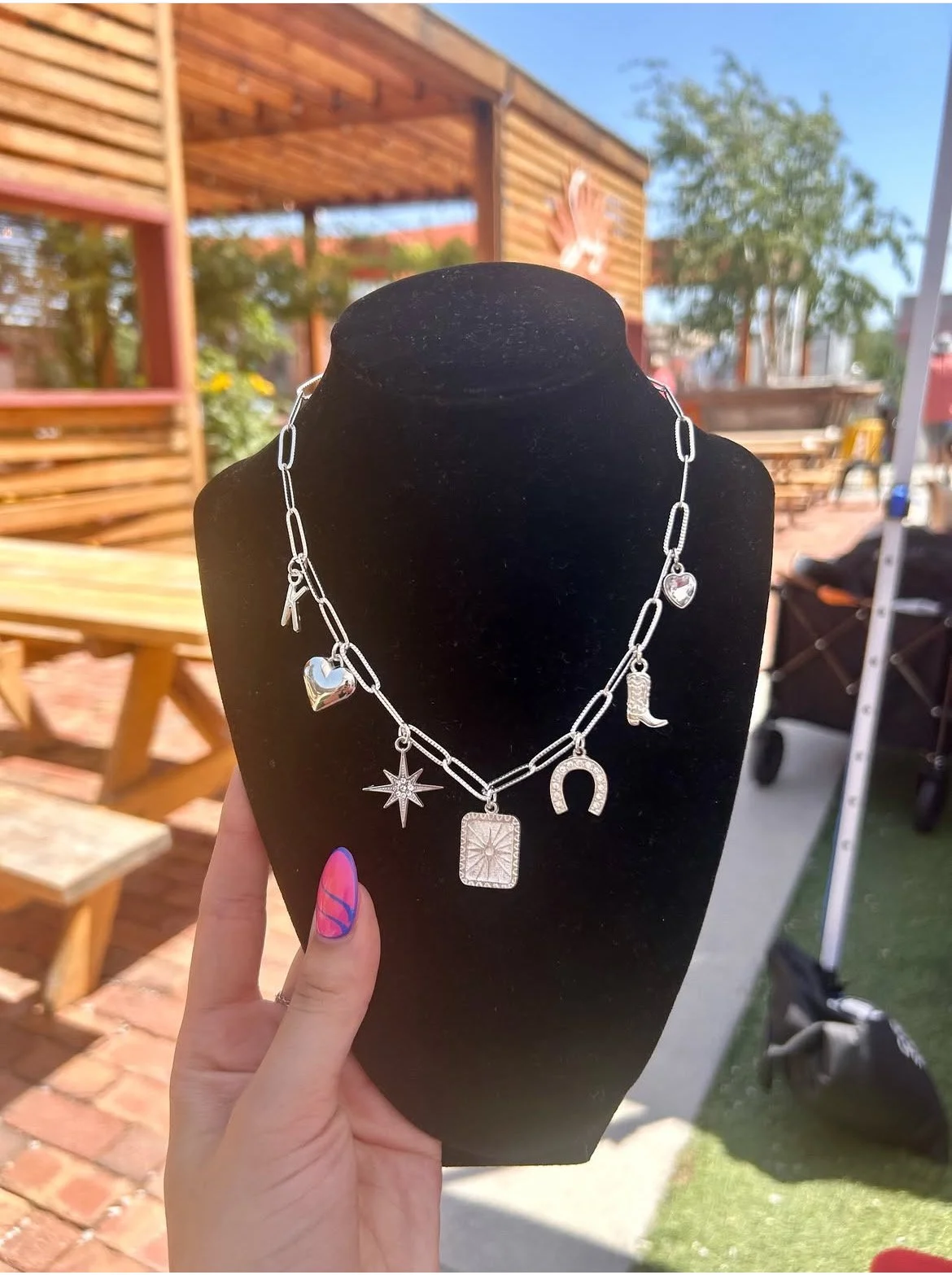 A silver charm bracelet with various charms, including a heart, star, rectangular pendant, horseshoe, and a lipstick, displayed on a black jewelry stand outdoors.