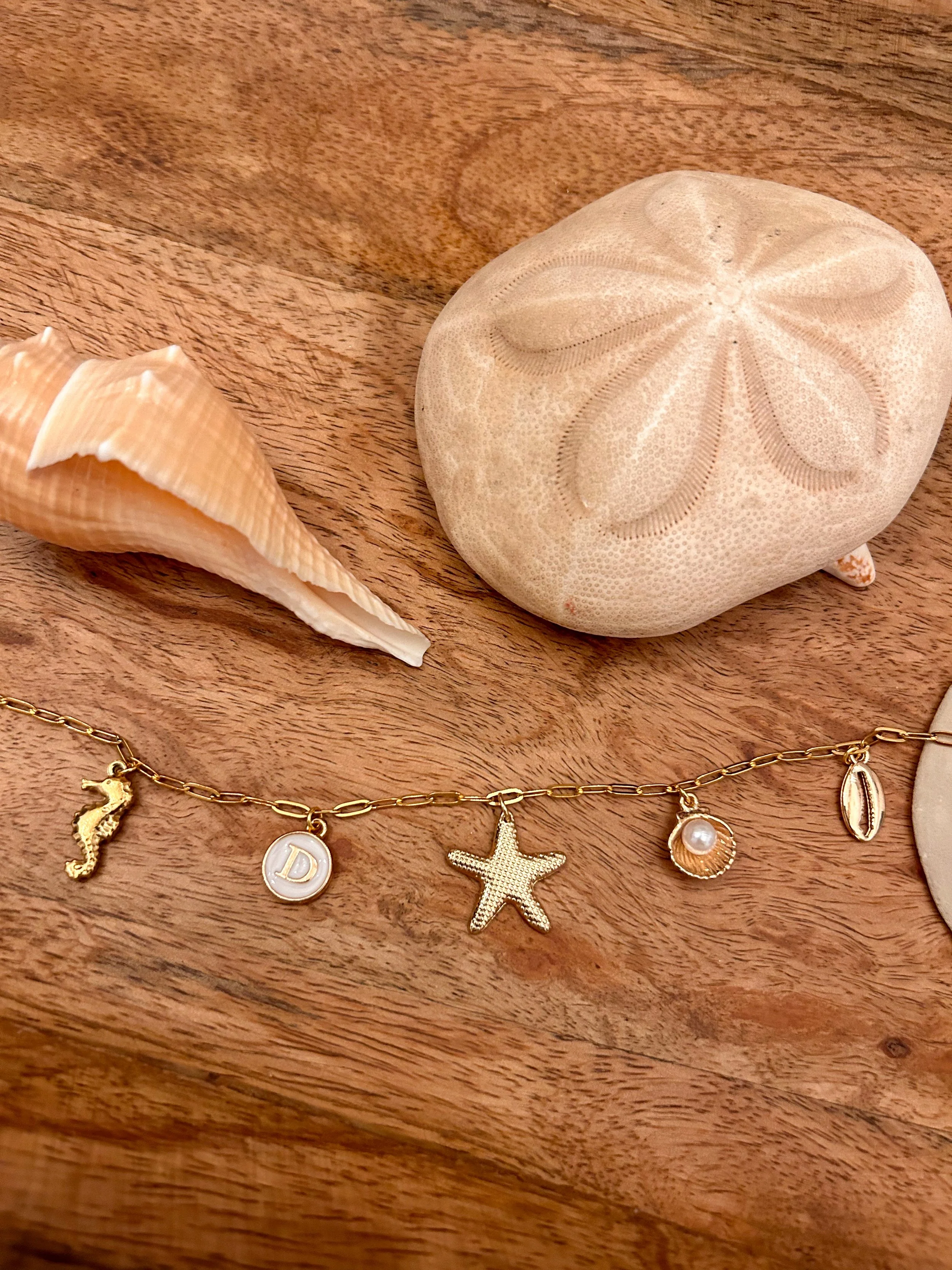 A bracelet with charms including a seahorse, a circular charm with the letter 'D', a starfish, a pearl in a shell, and a shell, lying on a wooden surface beside a seashell and a piece of coral.