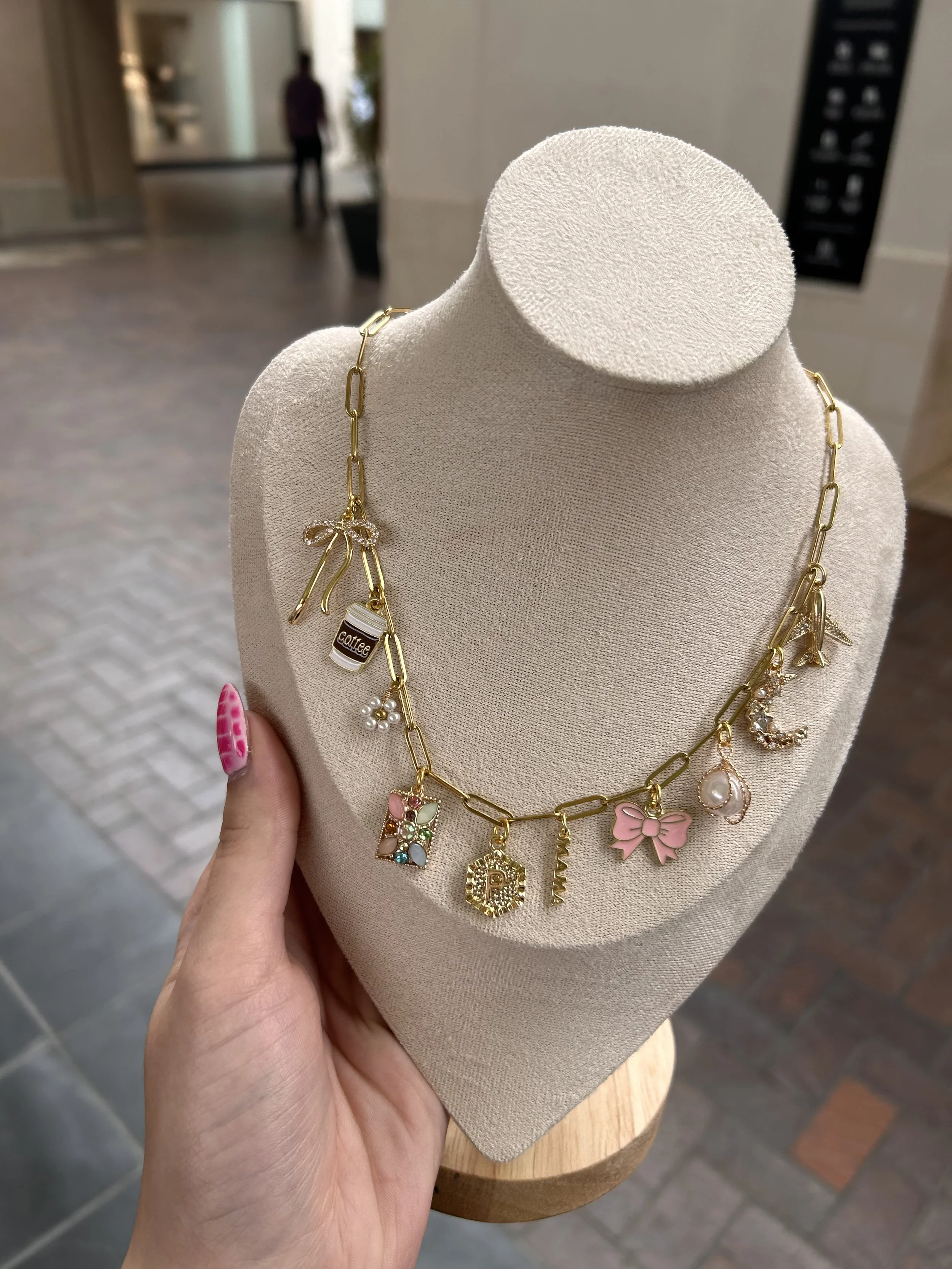 Gold charm necklace with various charms, including a coffee cup, pearl, flower, letter 'P', butterfly, and moon, displayed on a beige jewelry stand in an indoor setting.