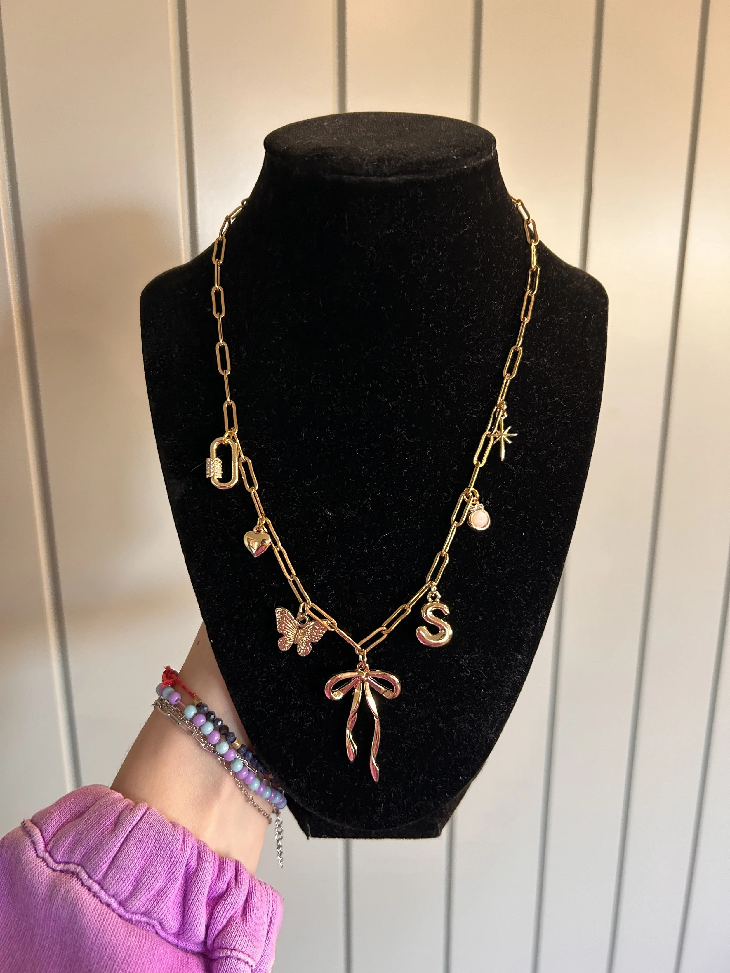 Gold chain necklace with various charms, including a bow, butterfly, letter S, and star, displayed on a black mannequin bust.