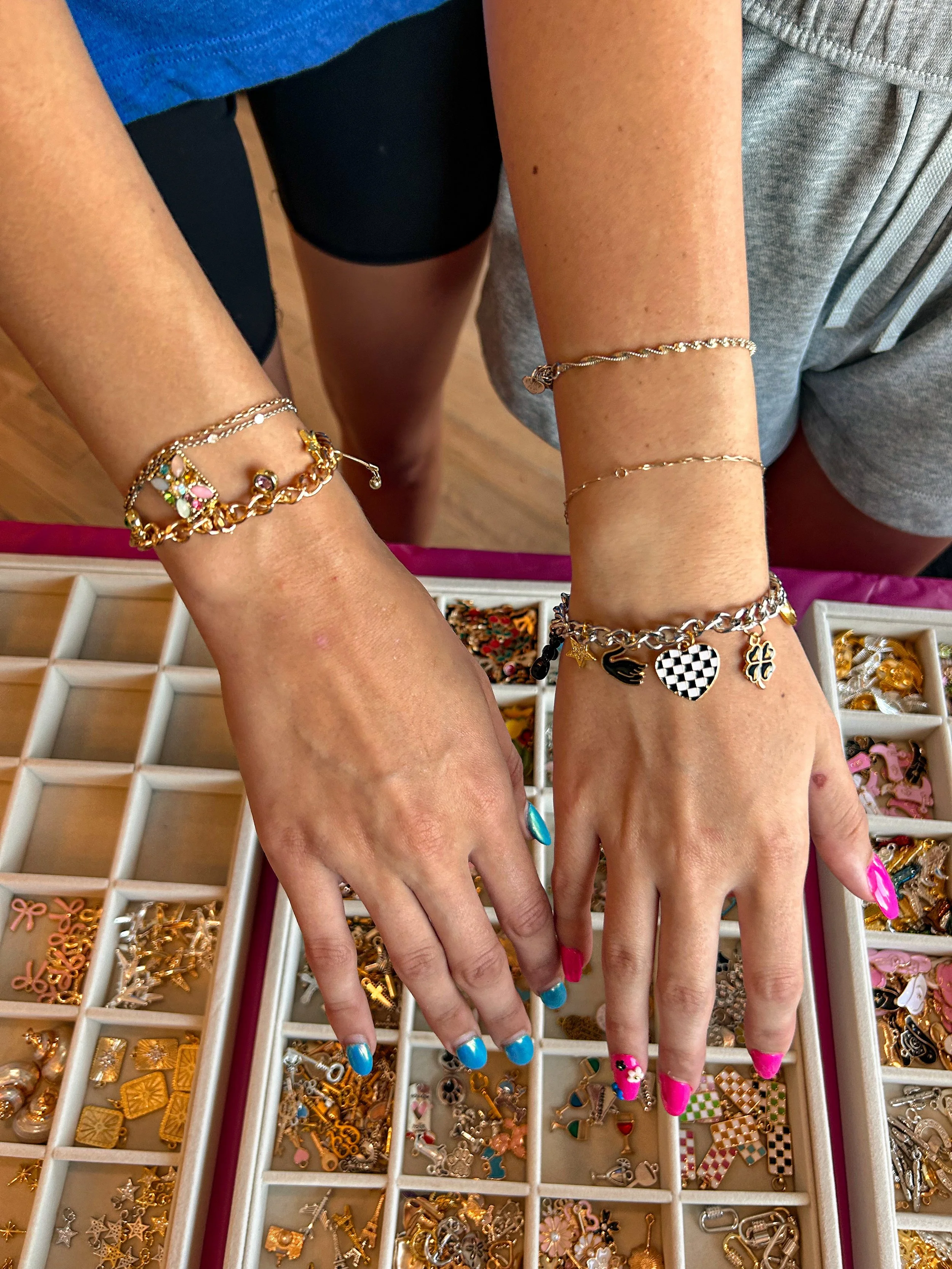 Two people display their hands with colorful nail polish, showing off various bracelets and rings, over a jewelry display tray filled with charms and jewelry pieces.
