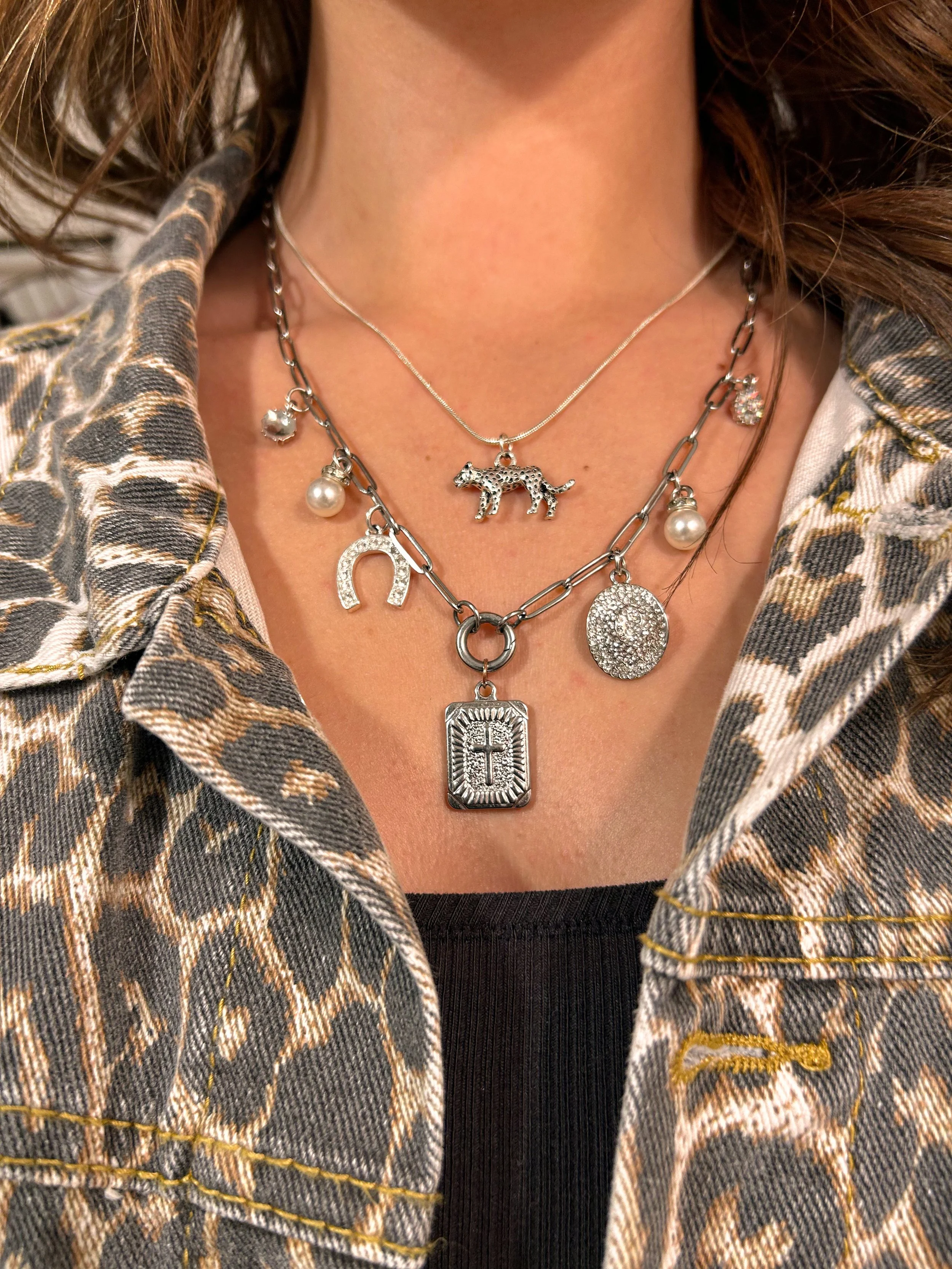 Close-up of layered necklaces with charms, including a leopard, horseshoe, pearl, sparkly stone, cross, and a round pendant, worn over a black top and animal print jacket.