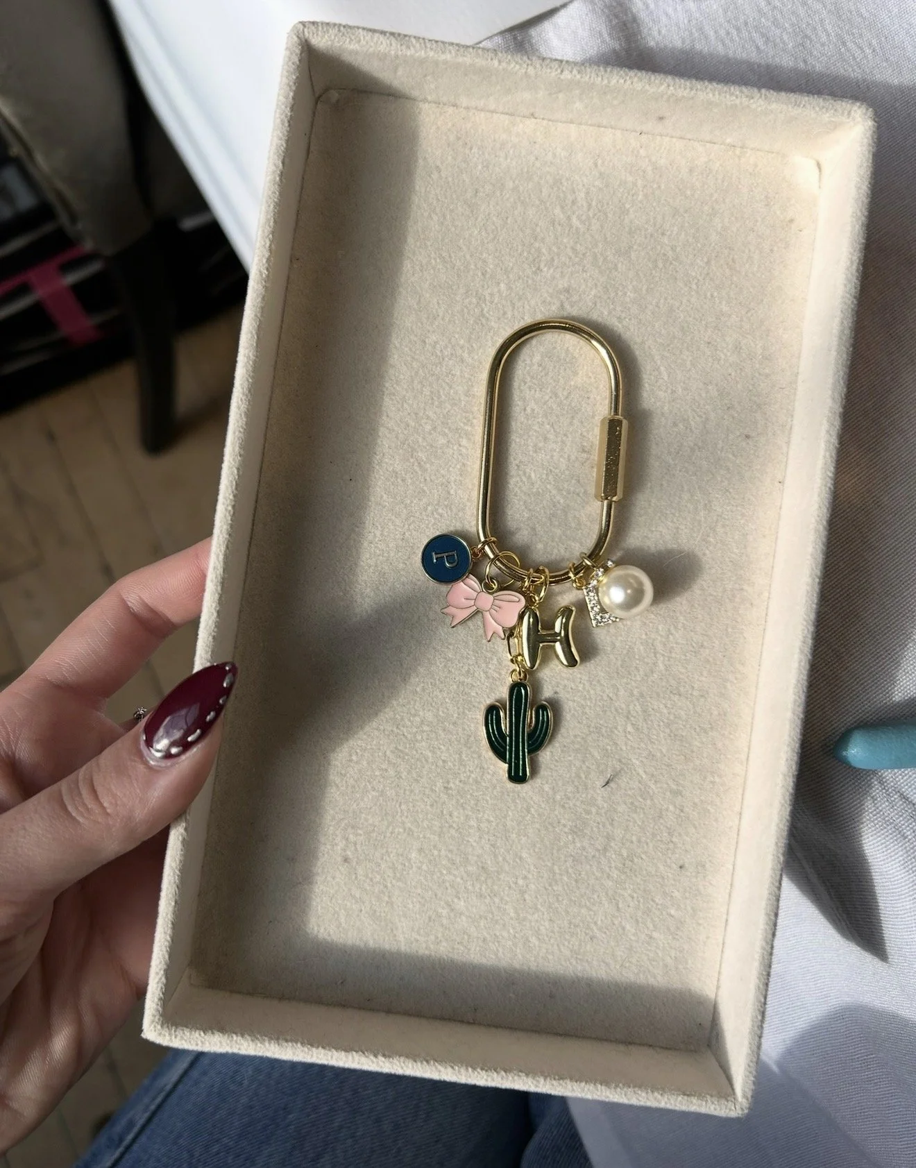 A gold bracelet with multiple charms, including a blue circle with a 'P', a pink bow, a pearl, a gold 'H', and a green cactus, inside a jewelry box.