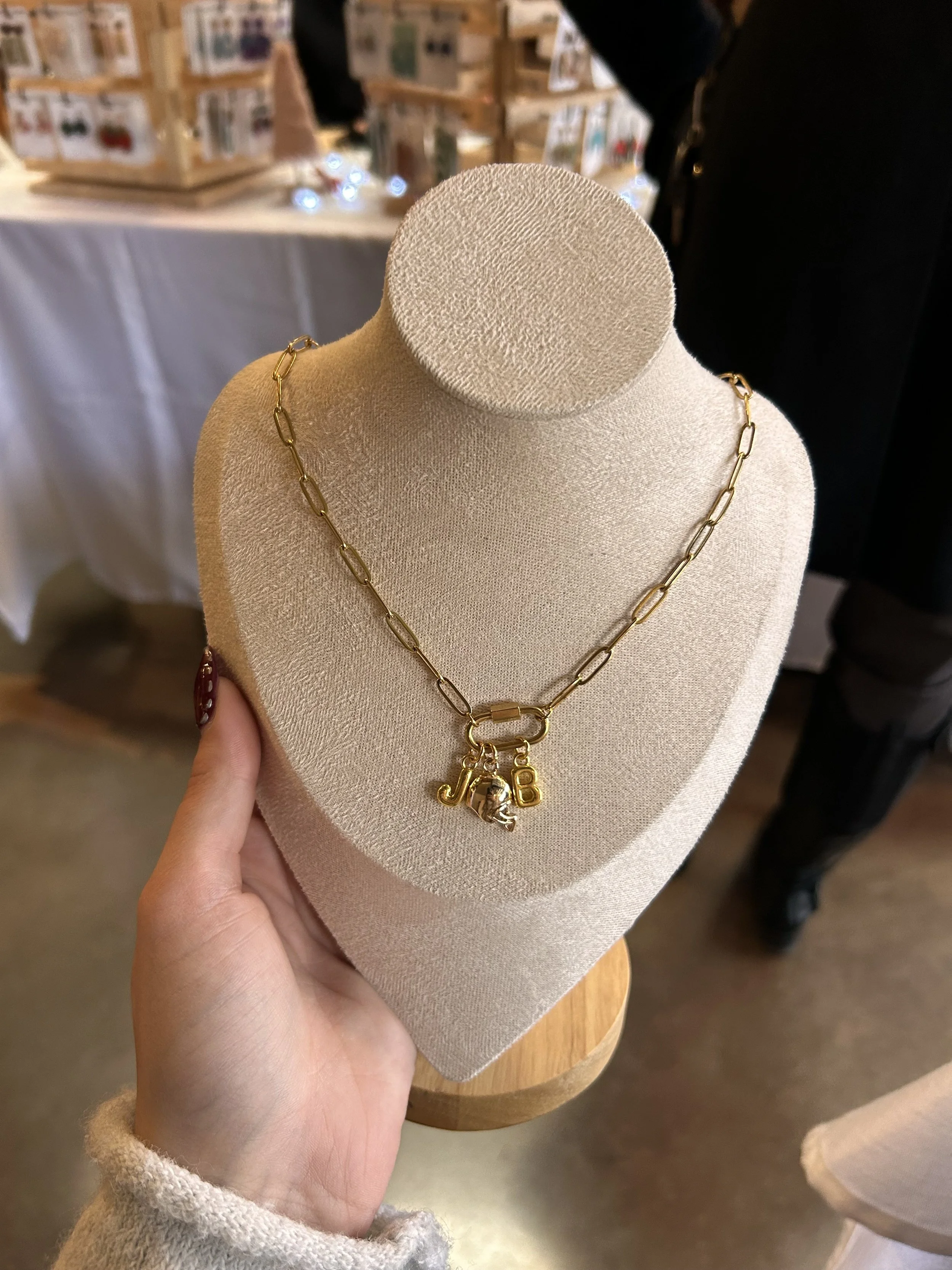 Gold-colored chain necklace with multiple charms, including a pink flower with a charm in the center, a round white gemstone, a pink cherry with green leaves, a pink circle with the letter 'S', and a gold heart, displayed on a black jewelry bust.