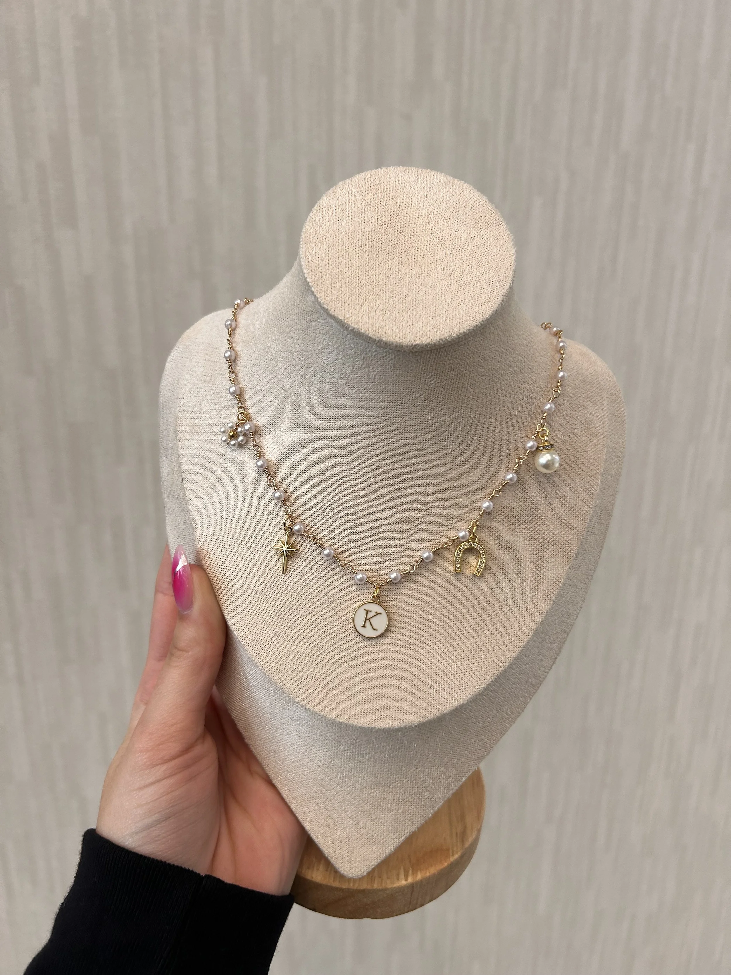 Necklace with pearl beads, gold charms including a star, horseshoe, and round pendant with the letter K, displayed on a beige jewelry bust.