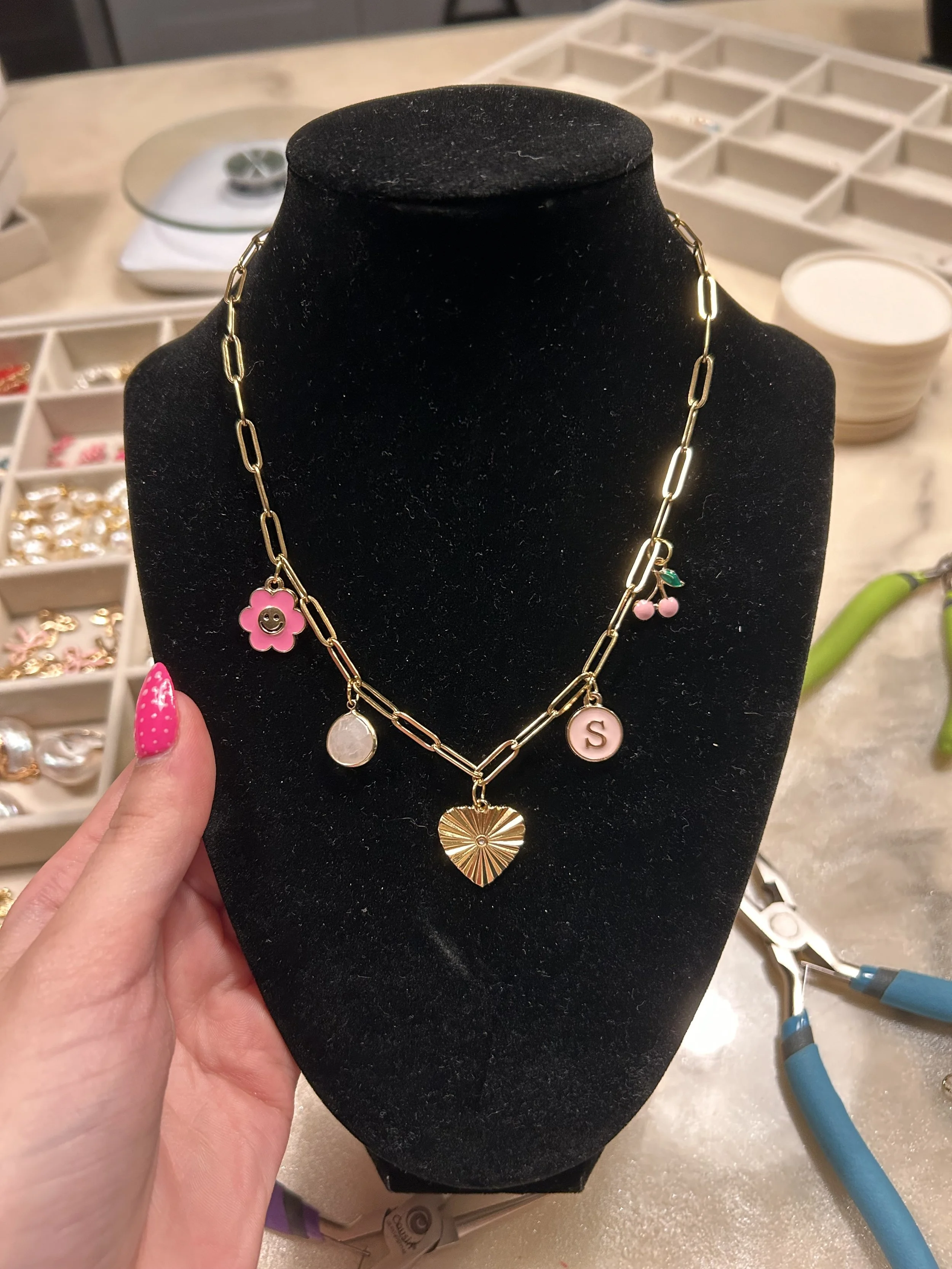 A black jewelry display bust showcasing a gold chain necklace with five charms, including a pink flower, a circular white pendant, a heart, a letter S, and a cherry charm with pink fruit and green leaves. The background shows organized jewelry trays 