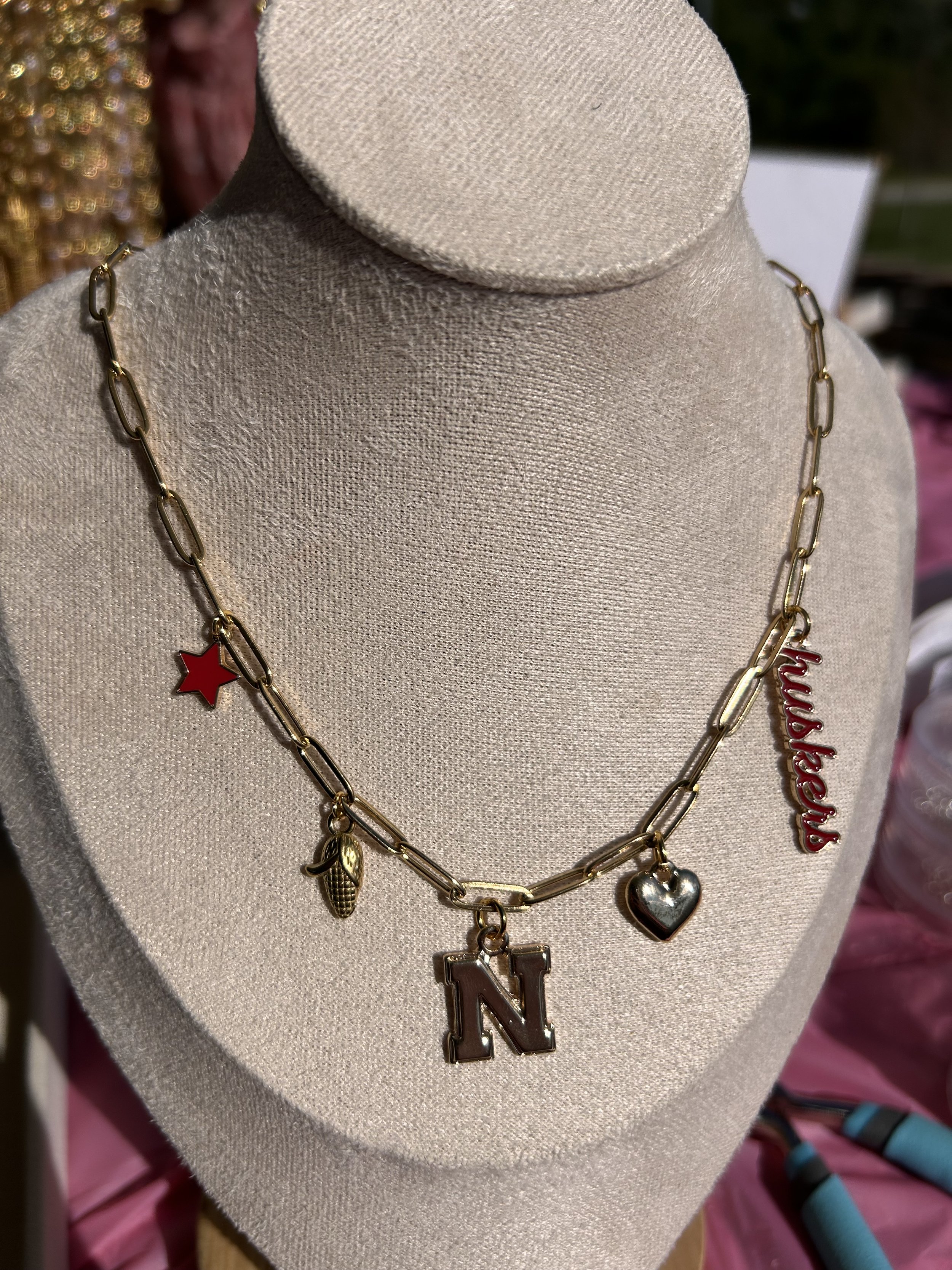 Gold-colored chain necklace with charms including a red star, a gold corn ear, a silver heart, a letter N, and the word "aughers" in red lettering.