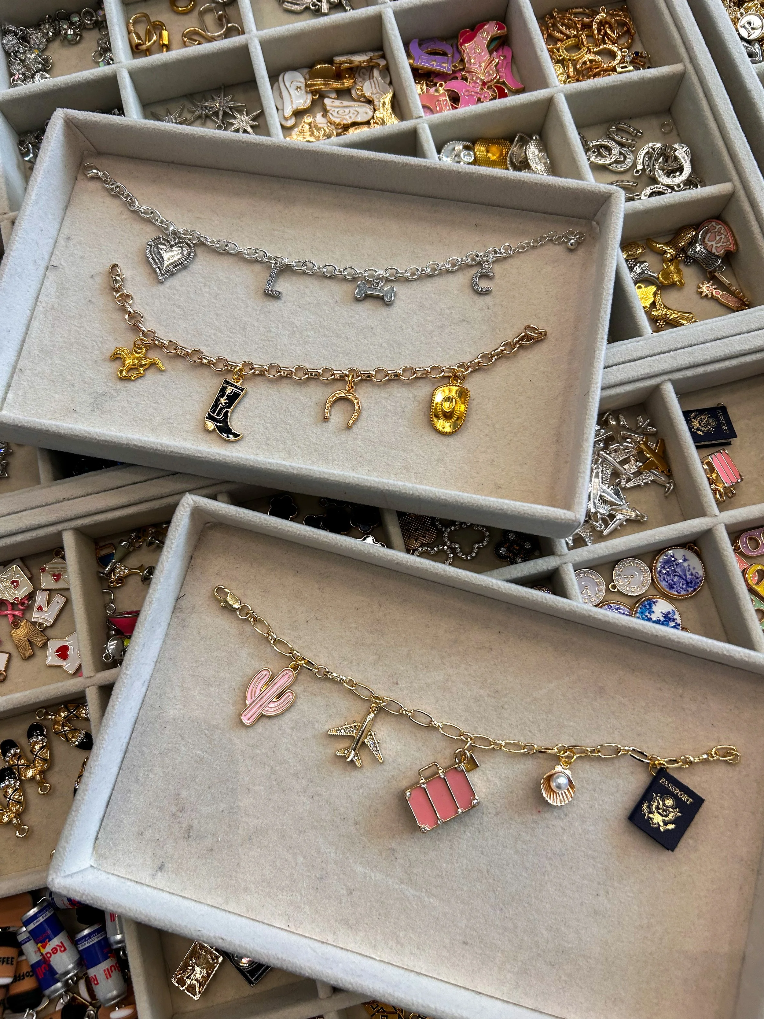 Two charm bracelets on display, one with silver and the other with gold, featuring charms like a heart, horseshoe, and a suitcase. Multiple jewelry boxes with various loose charms and jewelry pieces, including animals, stars, and decorative charms, are visible in the background.