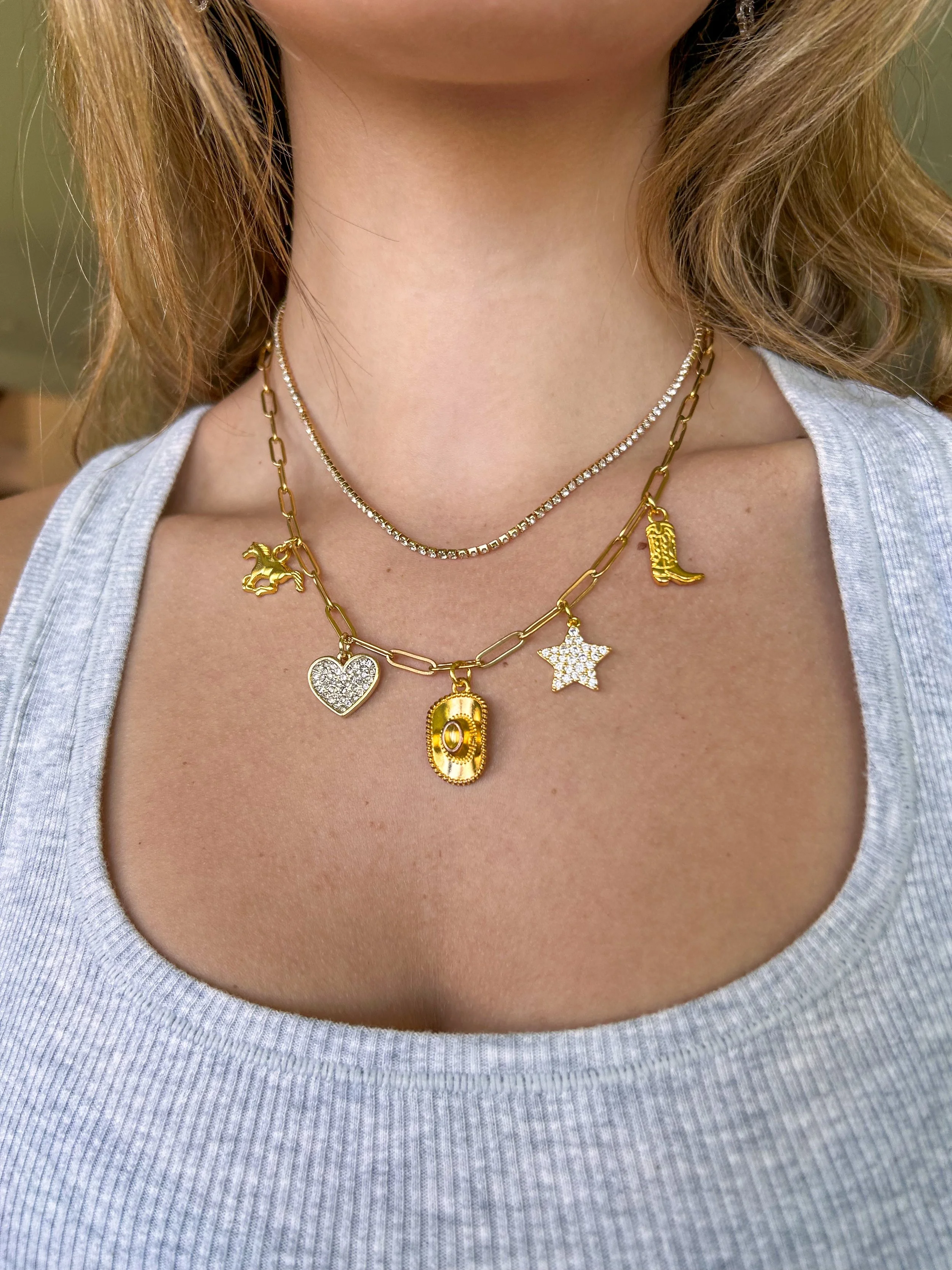 Close-up of a woman's neck and upper chest showing three layered gold necklaces with various charms, including a horse, a heart, a boot, a dollar sign, and a star, against a gray tank top.