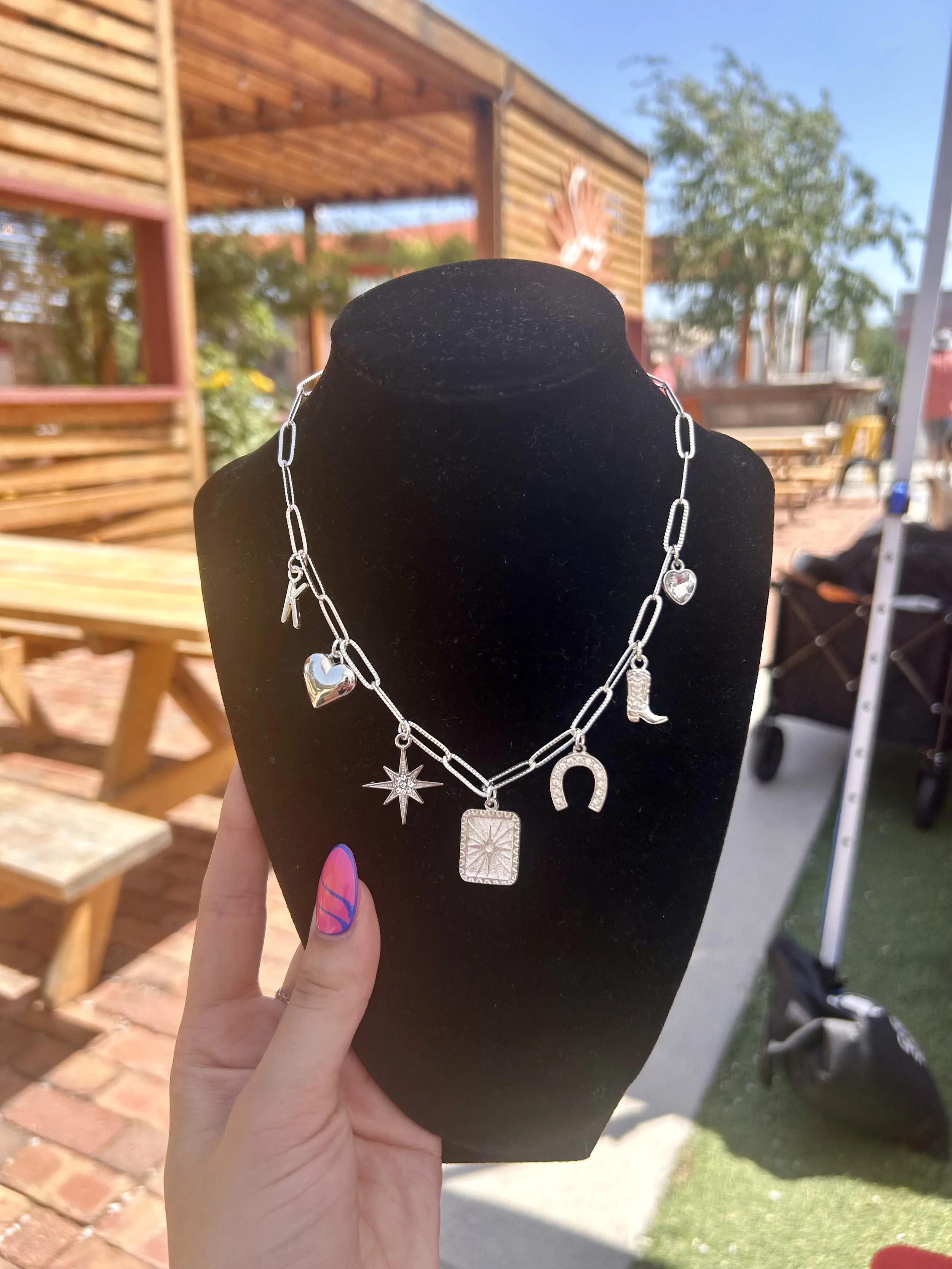 Silver necklace with various charms including hearts, a star, a horseshoe, and a rectangle, displayed on a black velvet bust outdoors.