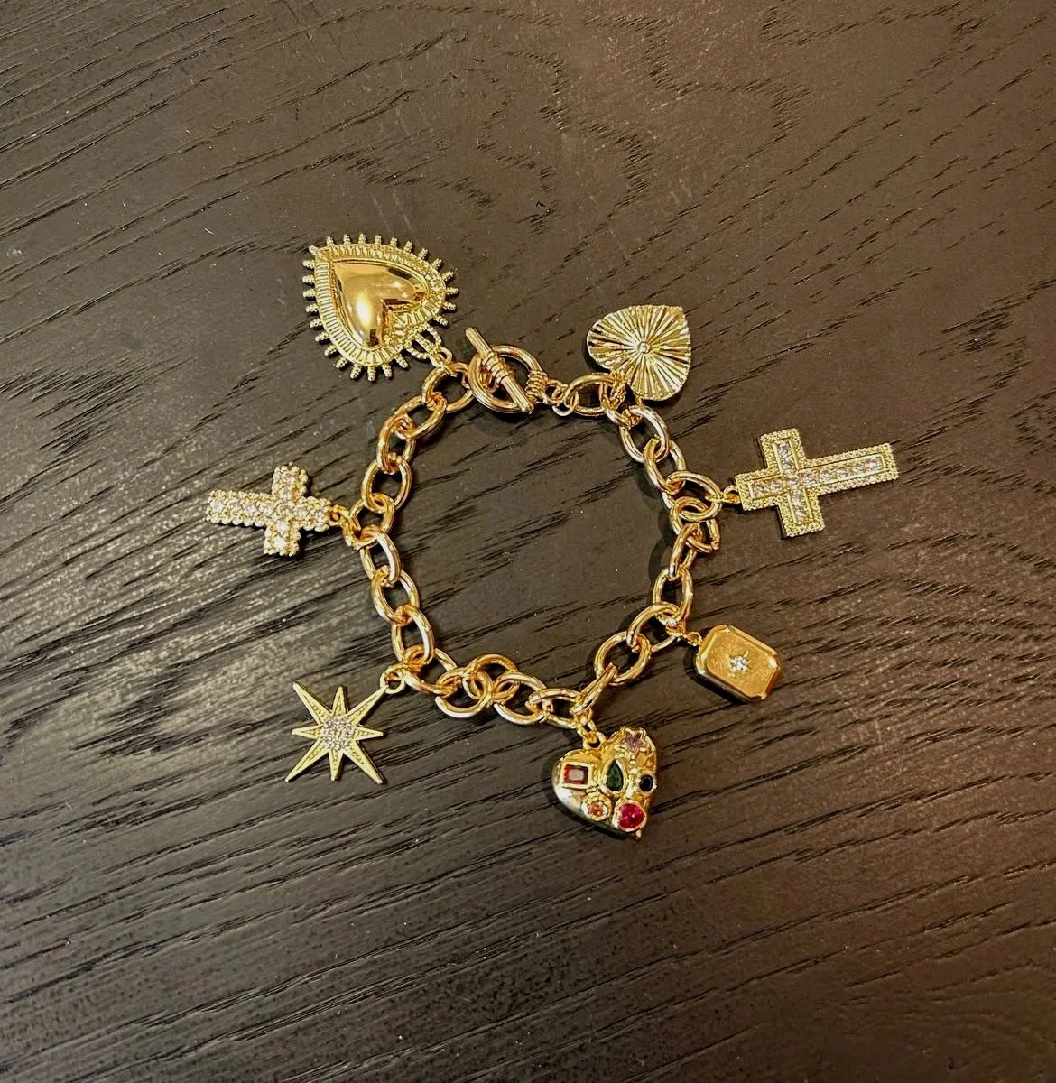 Gold chain bracelet with multiple decorative charms including a heart, cross, star, and colorful gemstone-encrusted pieces.