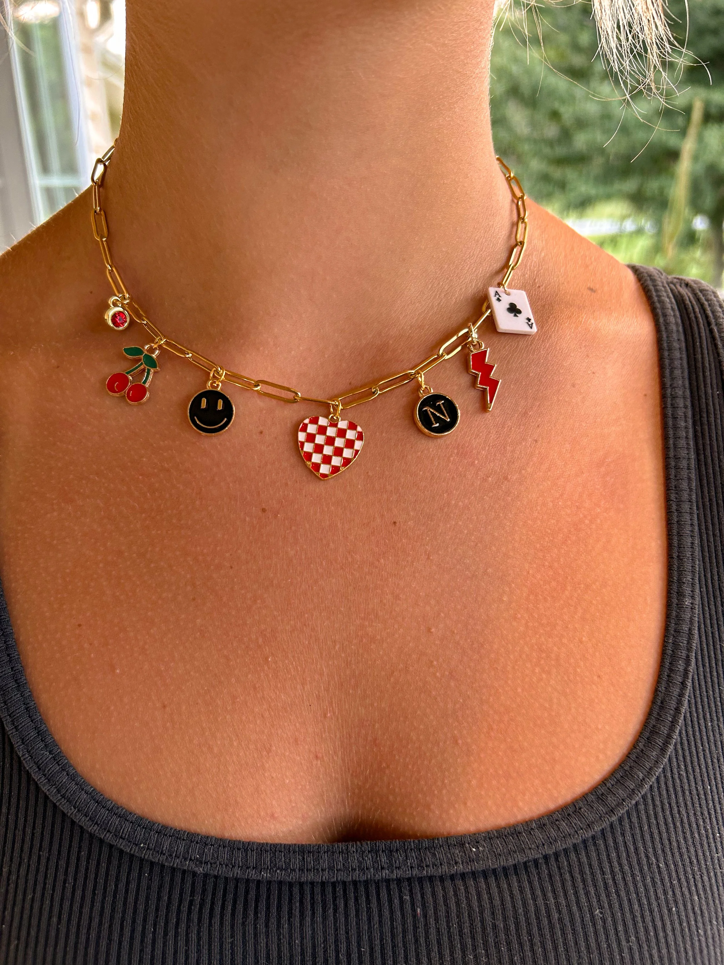 Close-up of a woman's neck wearing a gold chain necklace with various colorful charms, including cherries, a smiley face, a checkered heart, a 'N' letter charm, a lightning bolt, and a small white playing card with the ace of clubs, against a dark sh
