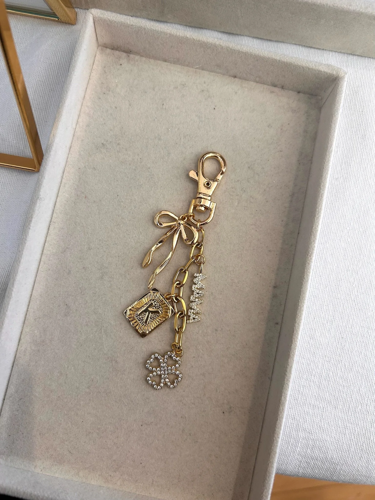 Gold keychain with multiple charms including a bow, a diamond-shaped charm, a word charm, and a four-leaf clover, inside a light-colored jewelry box.