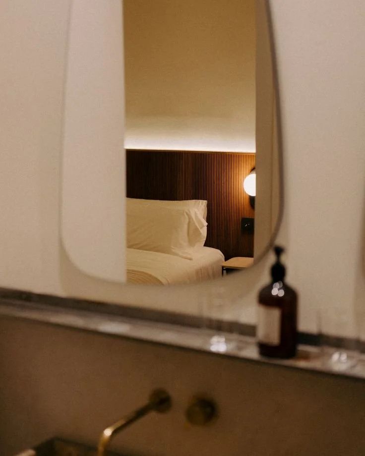A hotel room reflection seen through a mirror, showing a bed with white bedding and pillows, a dark wood headboard, and a bedside lamp emitting warm light.