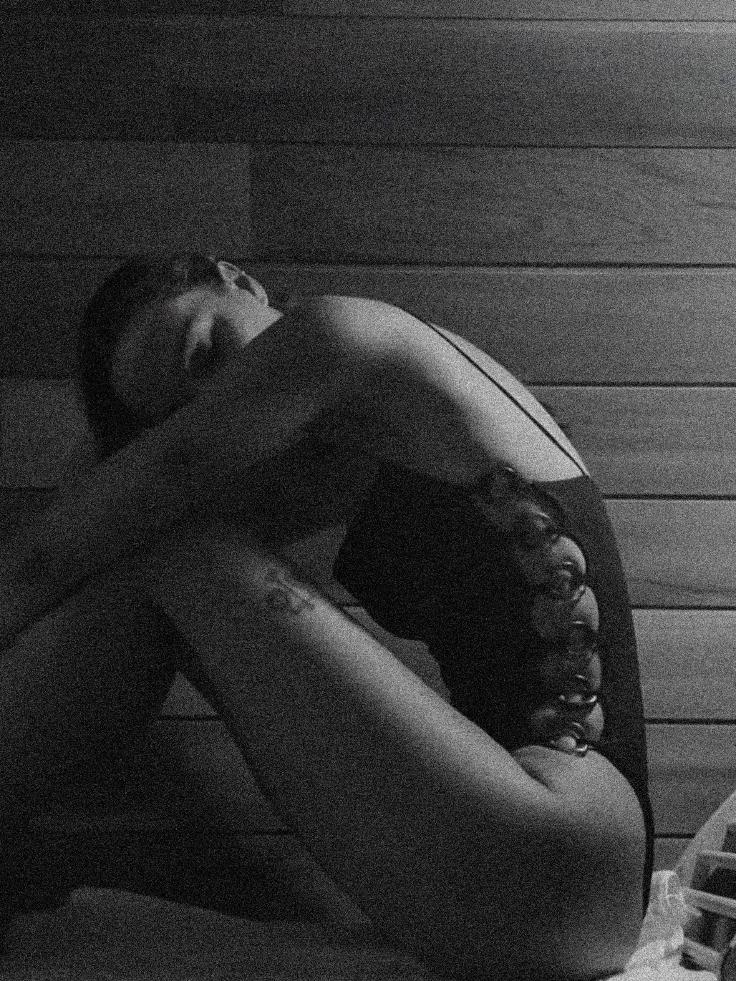 A woman sitting on the floor with knees pulled to chest, wearing a black dress, in front of a wooden wall.