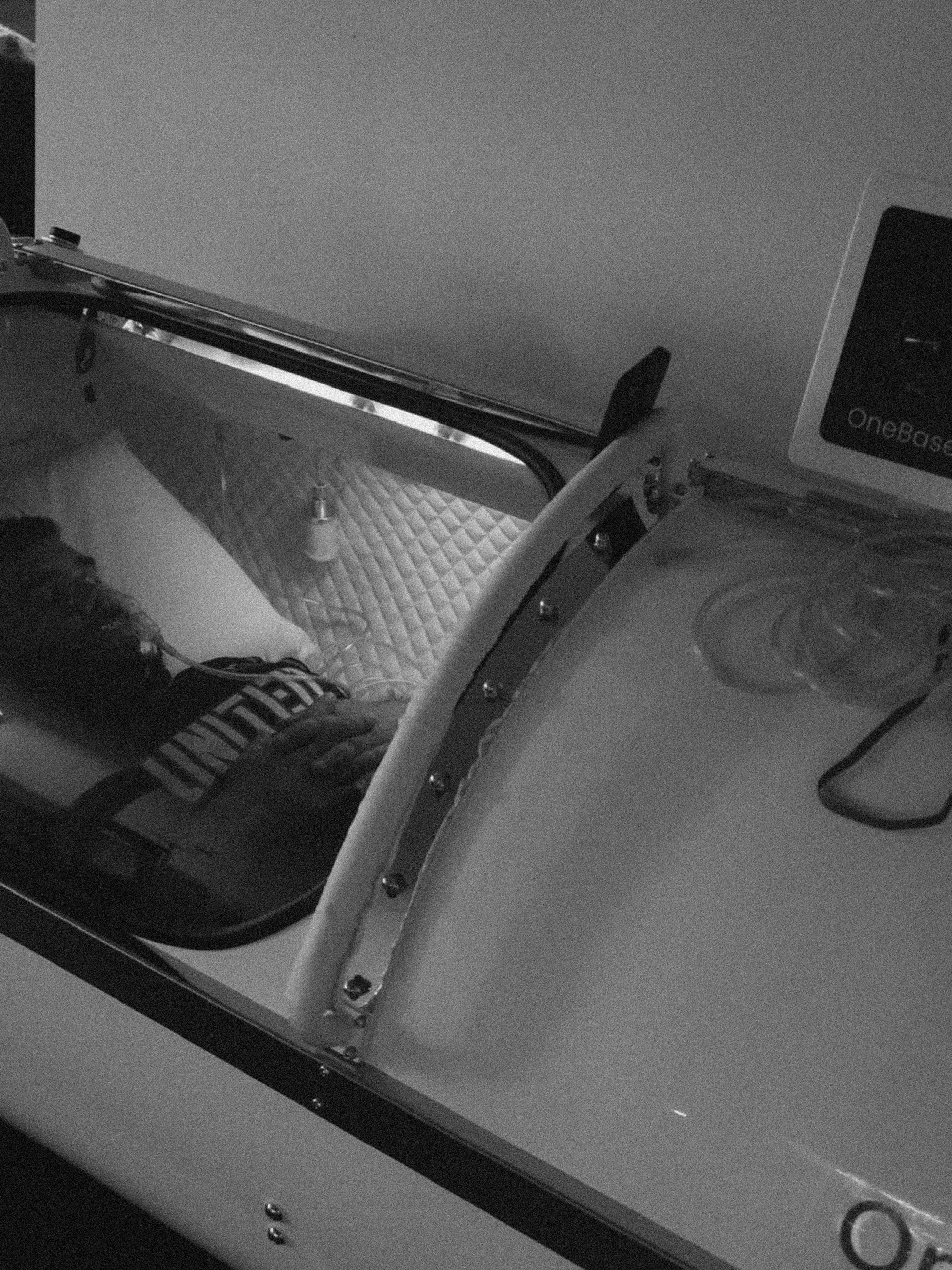 A patient lying on their back inside a medical hyperbaric chamber with a breathing mask, connected to medical equipment and a monitor.