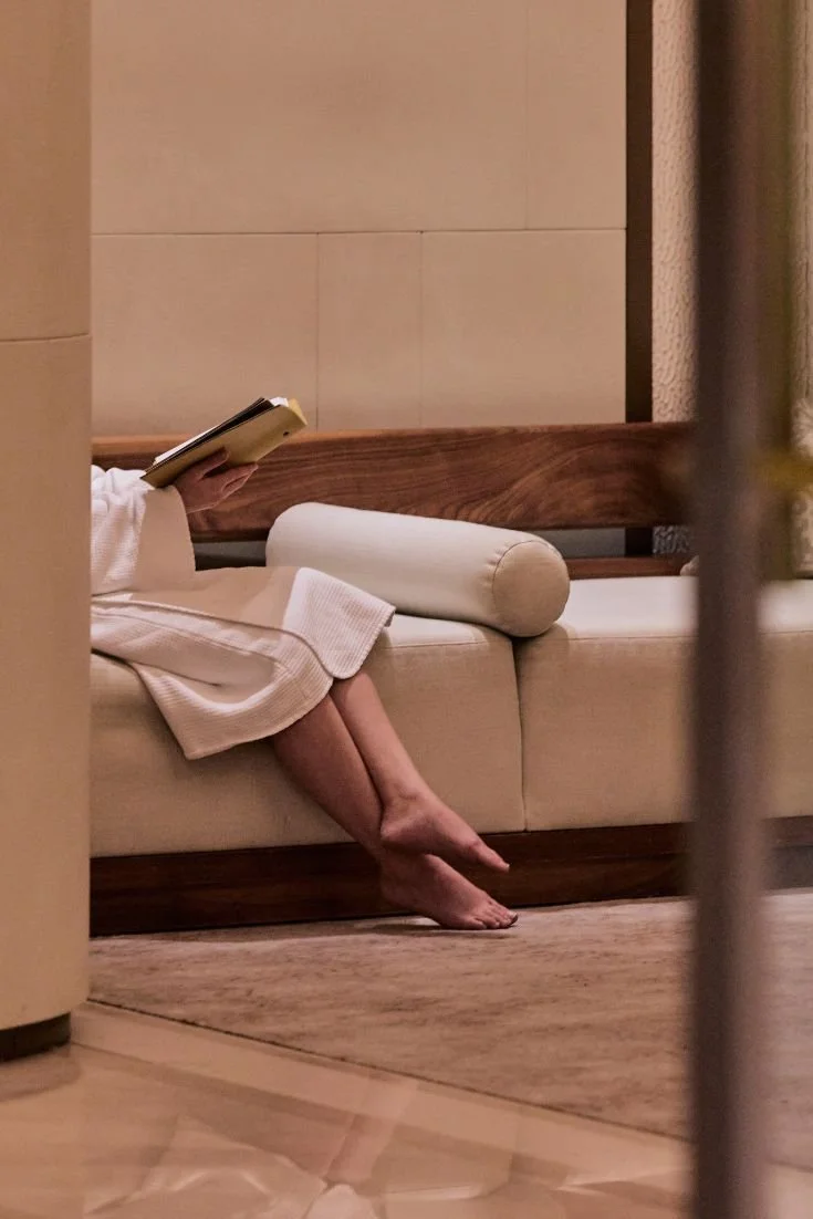 A person sitting on a beige sofa, reading a book, wearing a white robe, with their legs crossed at the ankles, and their feet on a wooden floor.