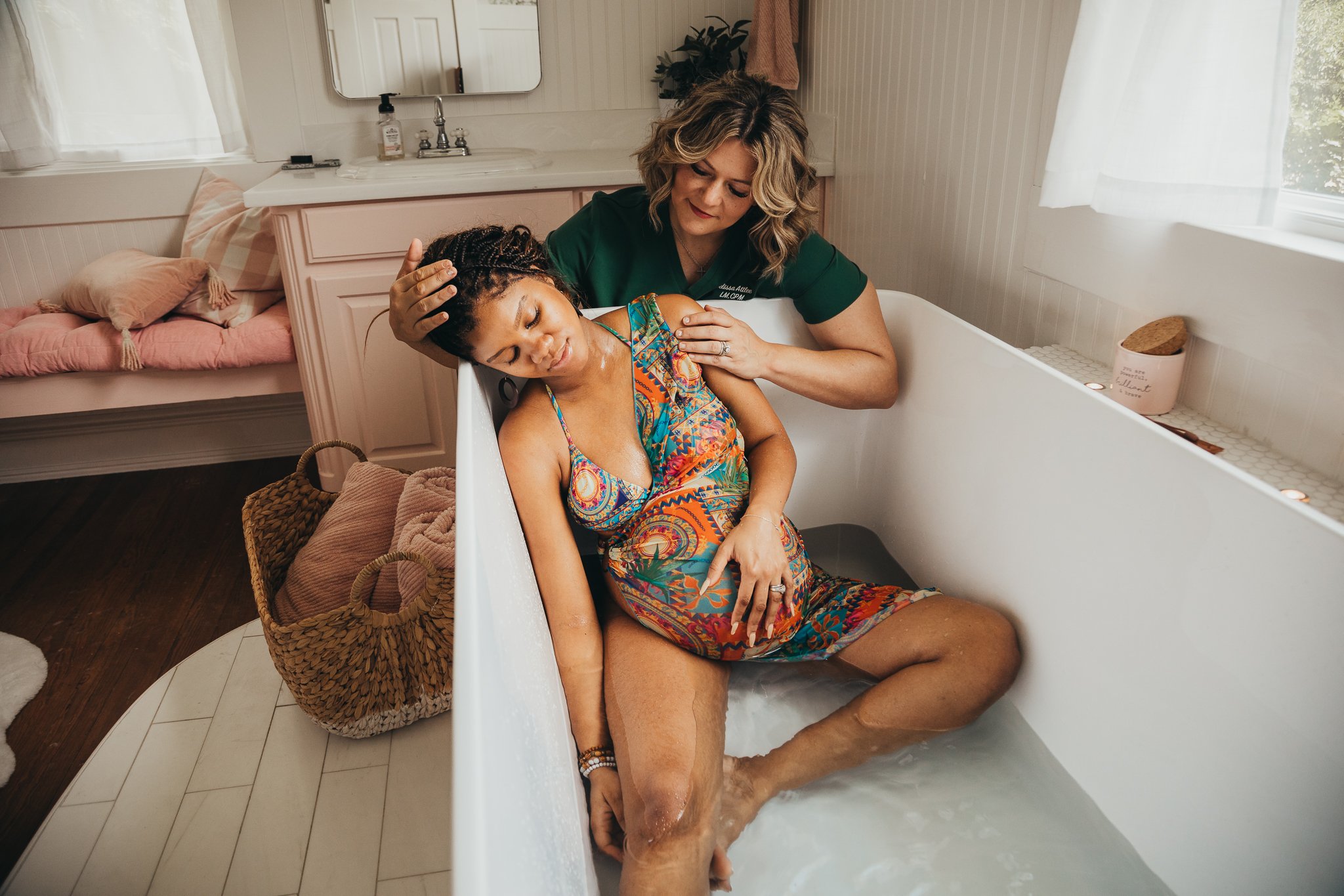 A woman in a colorful dress is sitting in a bathtub with water, with a caregiver supporting her head and shoulder in a gentle manner in a bright bathroom.