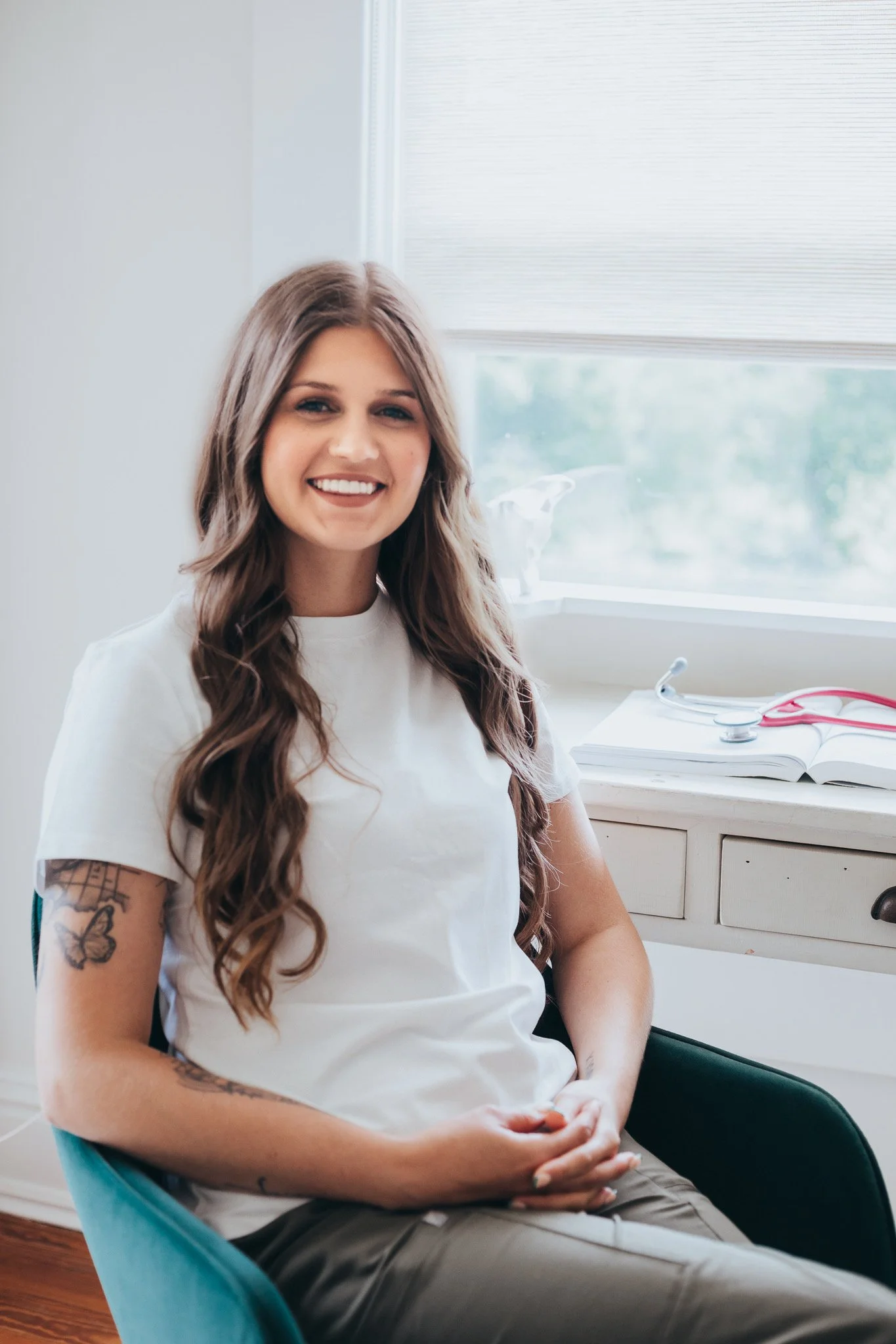 A young woman with long wavy brown hair, wearing a white t-shirt, sitting in a teal chair in a bright room with a window behind her. She is smiling and has tattoos on her arm, with a stethoscope and open book on the desk behind her.