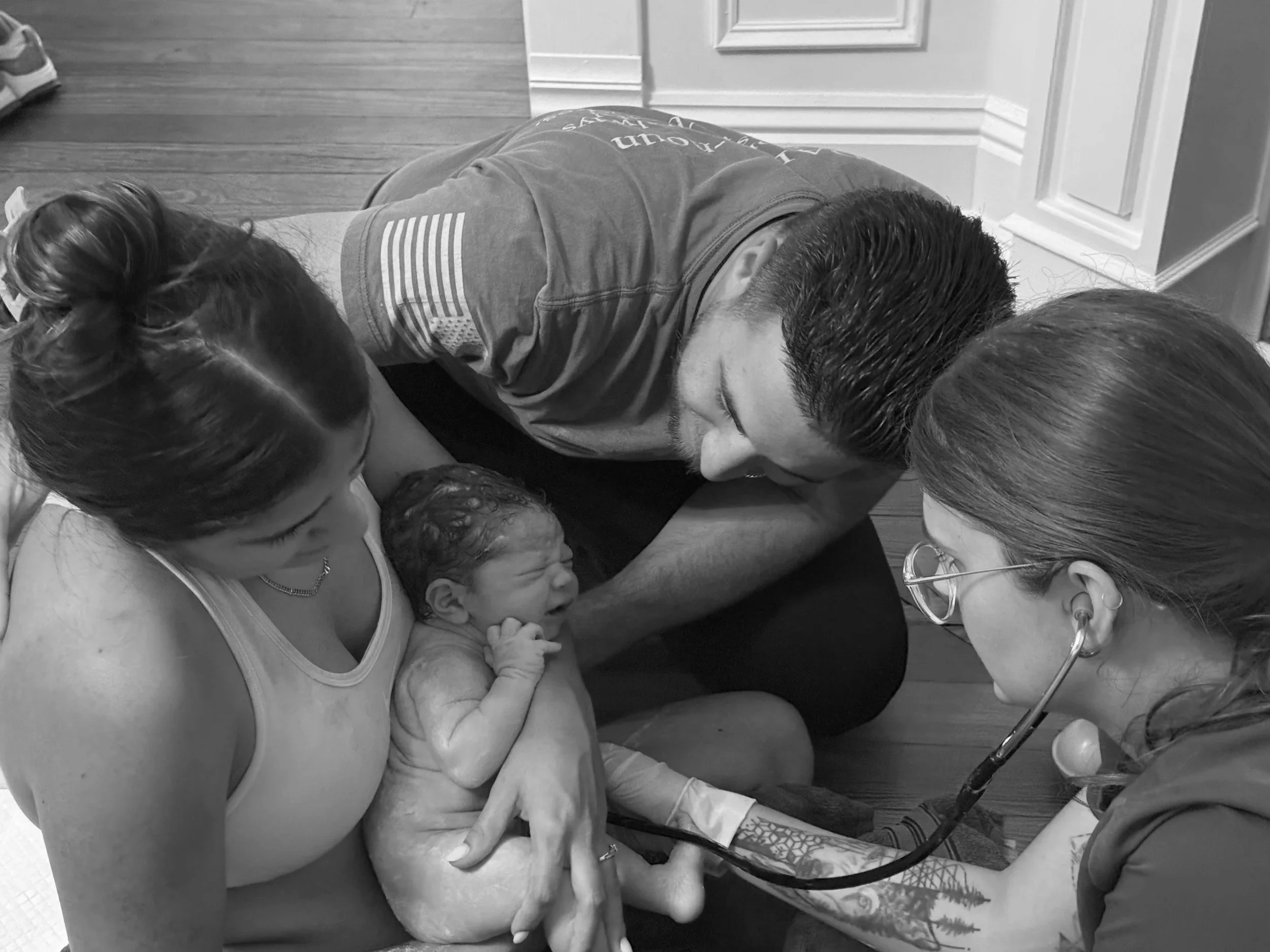 A woman holding a newborn baby during childbirth, with a man and a female healthcare professional with a stethoscope attending the baby in a home setting.
