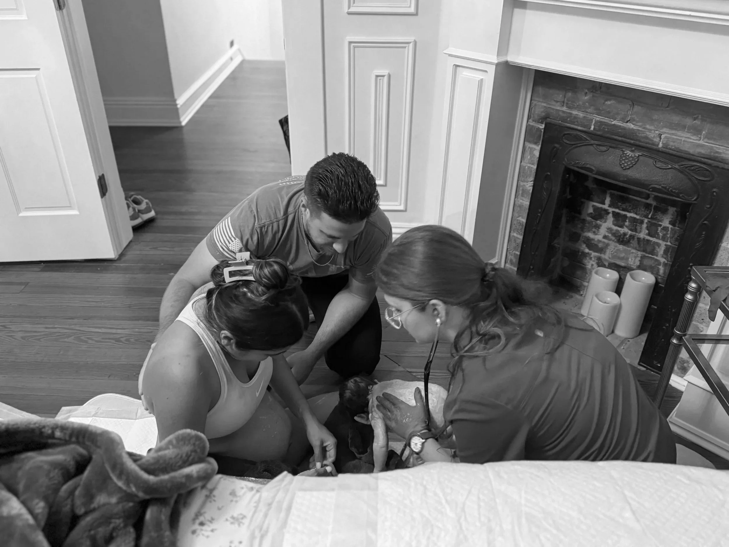 A woman in labor receiving medical attention from a paramedic and a man, with the woman holding a newborn baby, in a living room next to a fireplace.