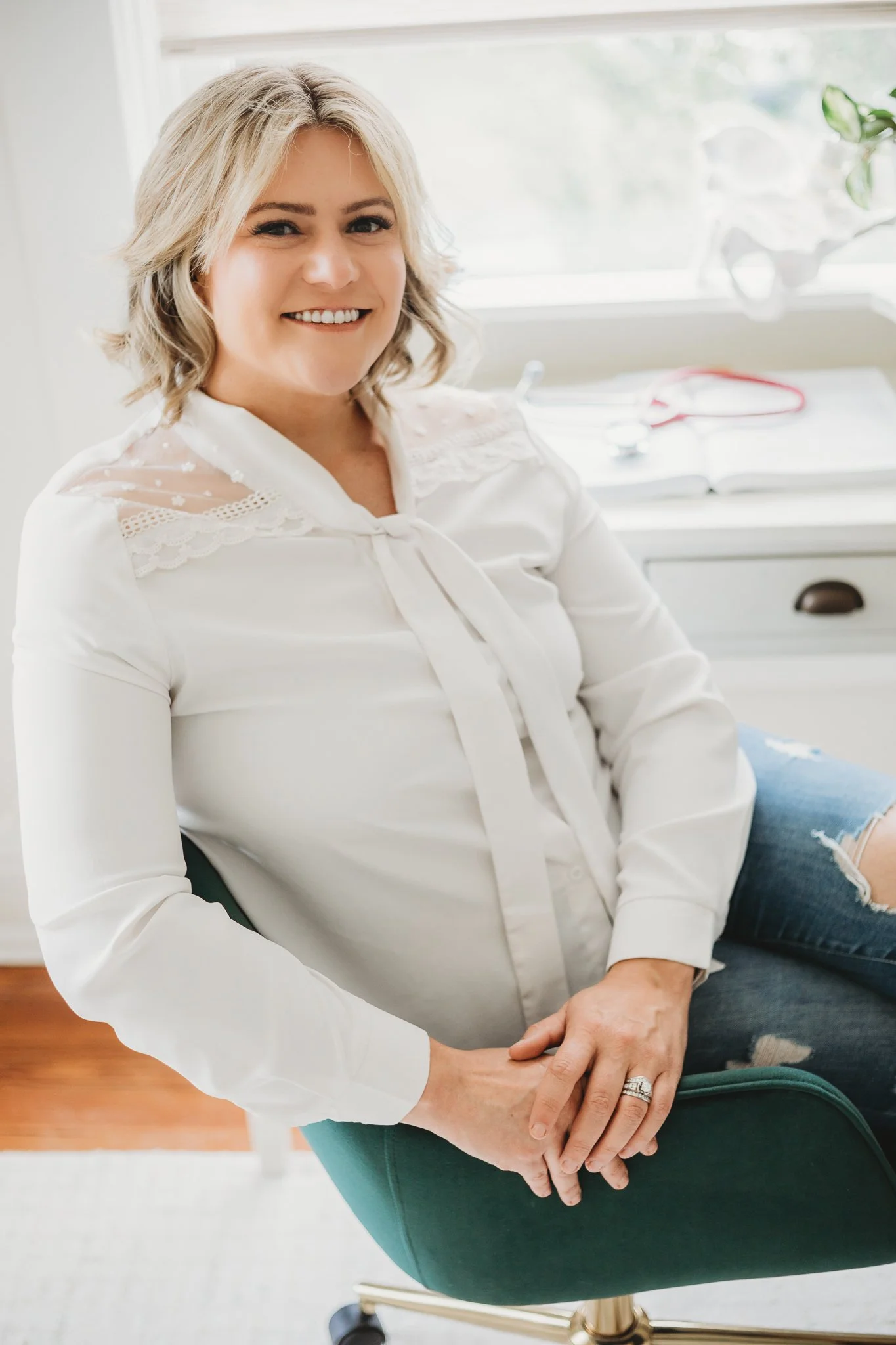 A woman with blonde hair smiling, sitting in a green office chair. She is wearing a white blouse and ripped jeans, with her hands resting on her lap. There is a white desk with an open notebook and a plant visible in the background.