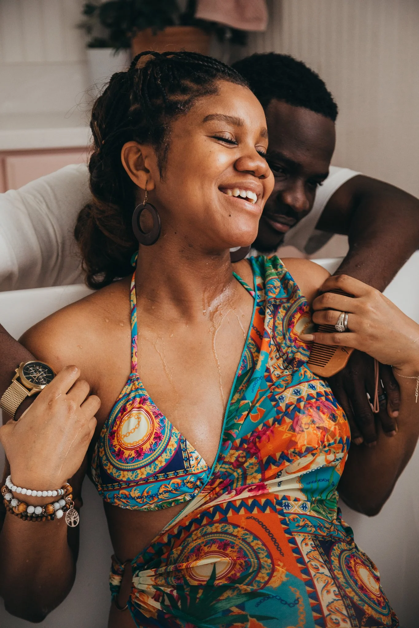A woman with wet skin and a colorful dress is smiling with her eyes closed, while a man behind her is combing her hair. They are in a bathroom, and she is wearing jewelry including earrings, bracelets, and rings.