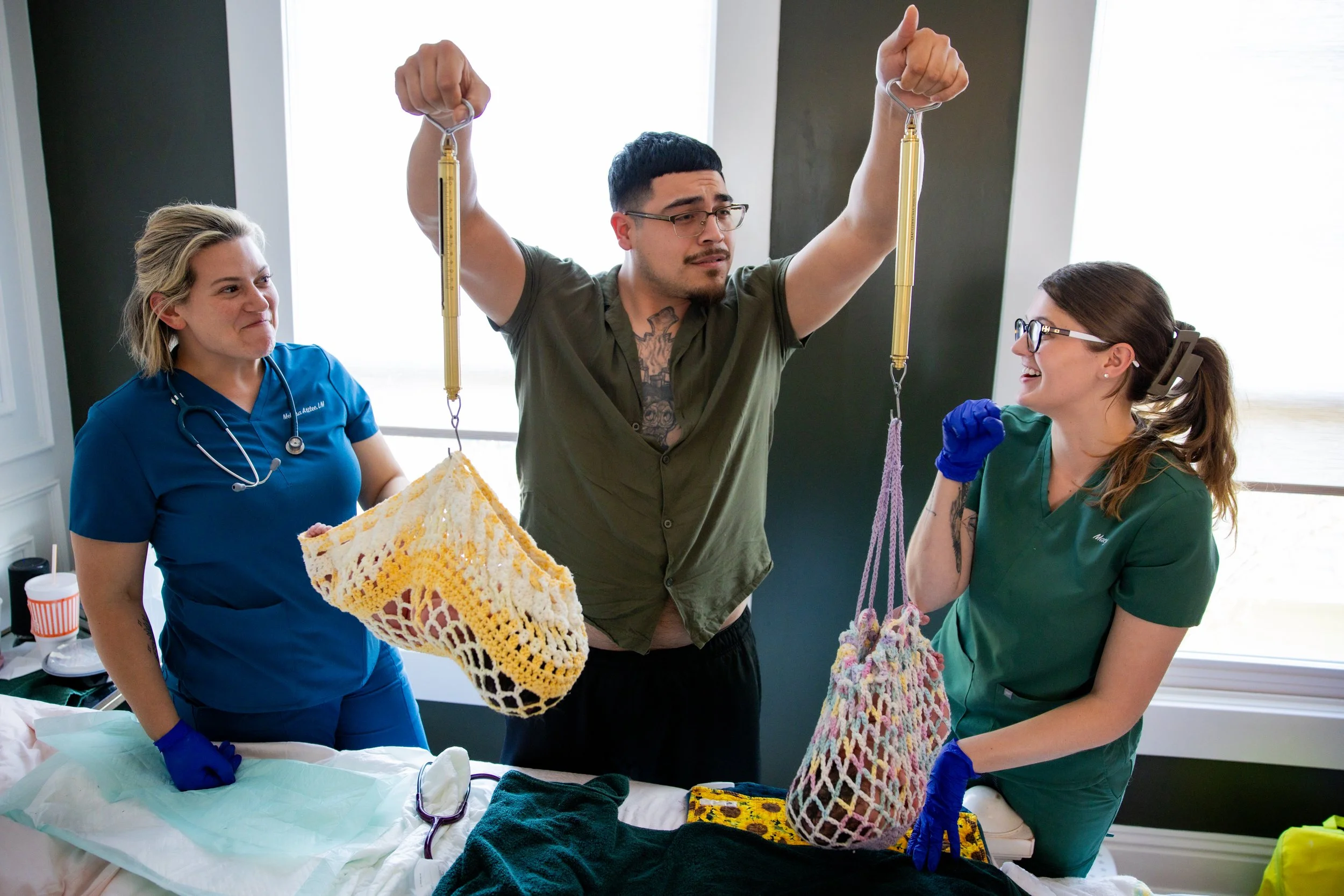 A man and two women, likely healthcare workers, are holding knitted stockings filled with an unknown substance, possibly demonstrating or explaining something in a medical setting. The women are wearing scrubs and gloves, and the man appears to be tr