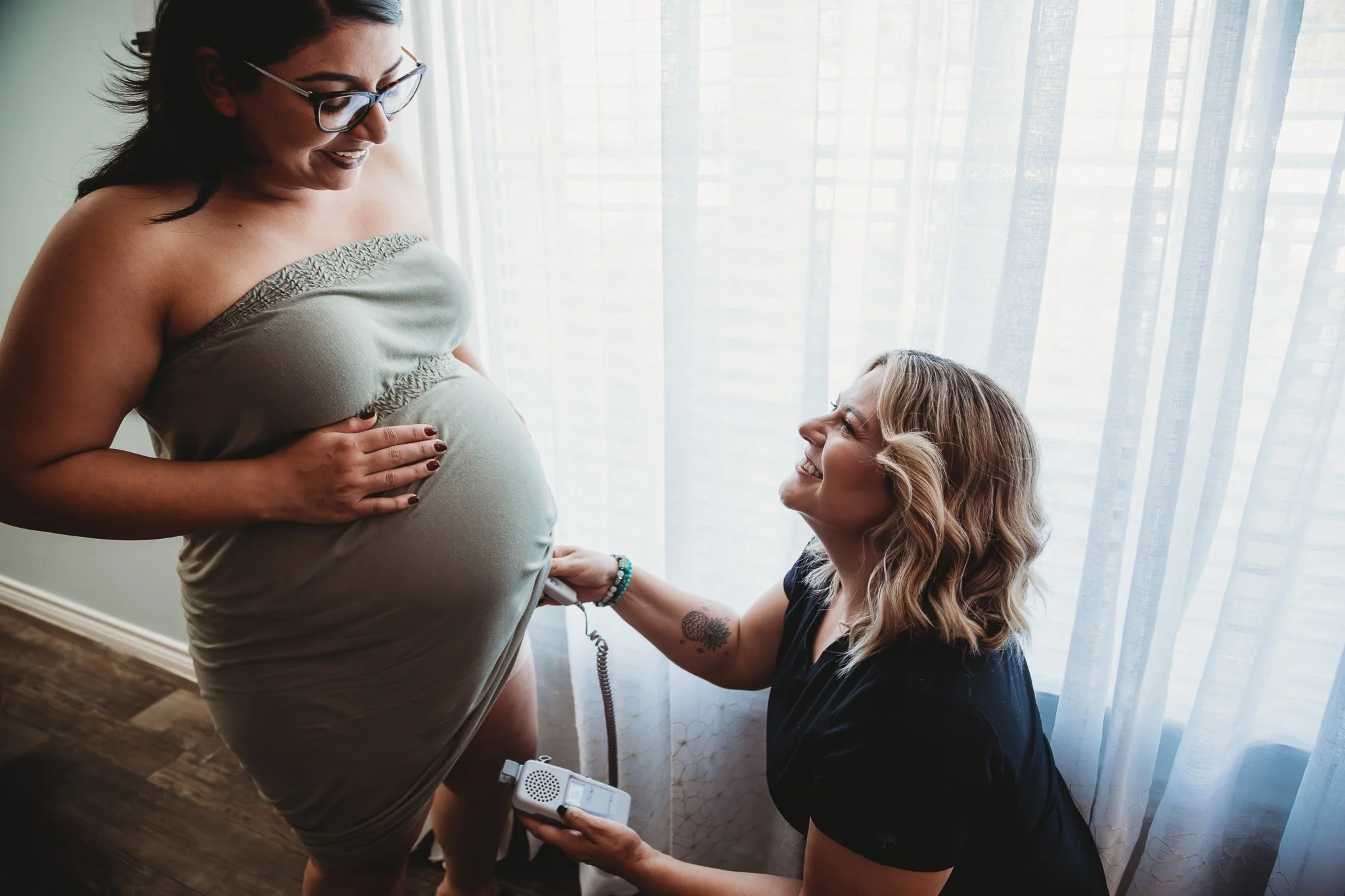 Pregnant woman smiling at her partner during a pregnancy ultrasound appointment, with a woman sitting on the floor holding ultrasound equipment and string.