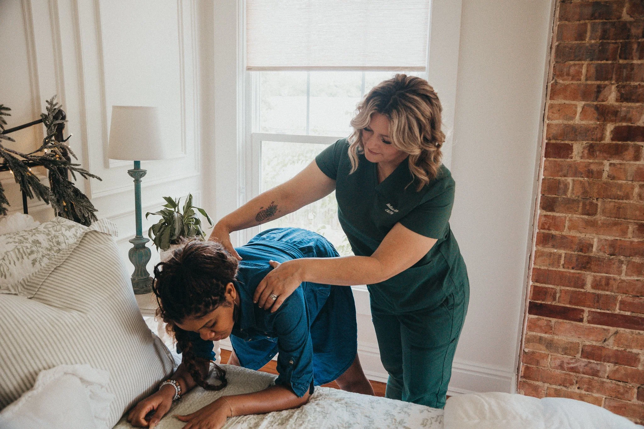 A woman receiving a massage from a massage therapist in a cozy bedroom with a brick wall, window, and plants.