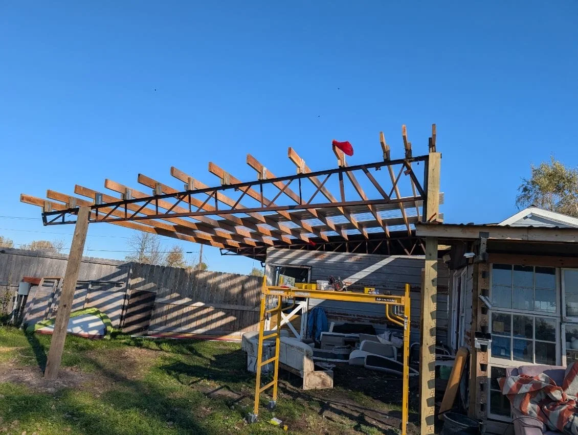 Under construction outdoor pergola with wooden beams and metal supports, yellow scaffolding, outdoor furniture, and a building with large windows under a clear blue sky.